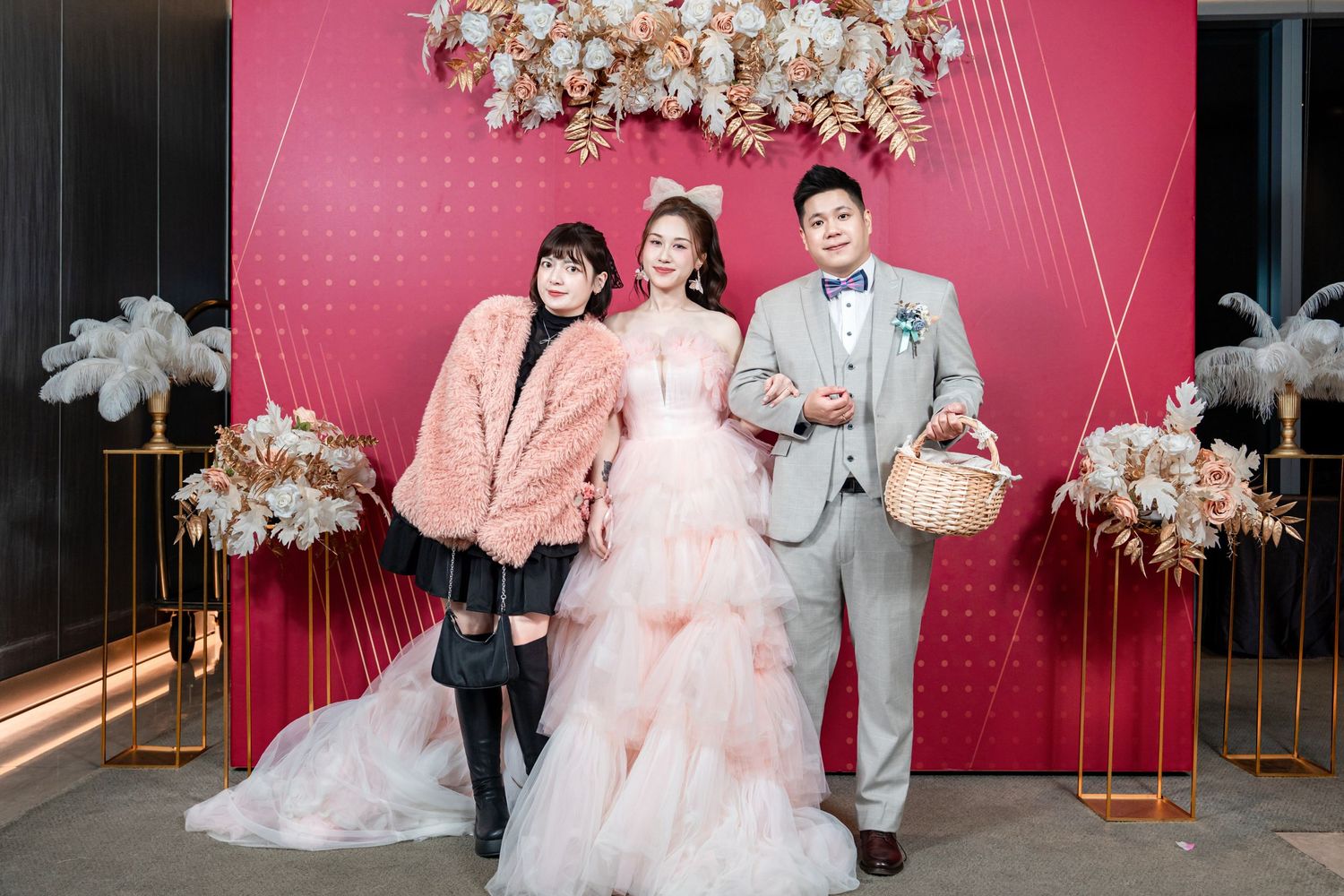 Three people pose together against a pink backdrop with floral decorations and a crystal chandelier at a formal event.
