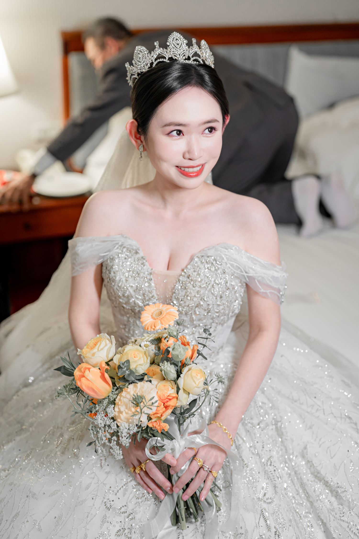 A bride in a sparkly white wedding gown holds a peach and orange floral bouquet against a light background.