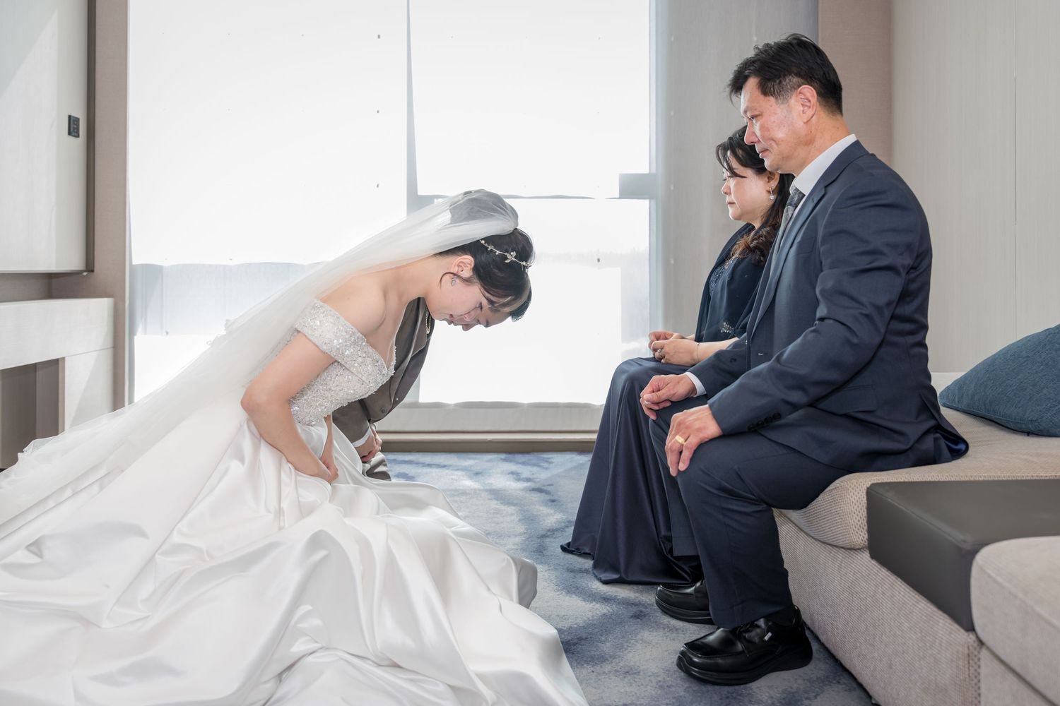 A bride in a flowing white wedding dress sits on modern gray steps in an elegant indoor setting.