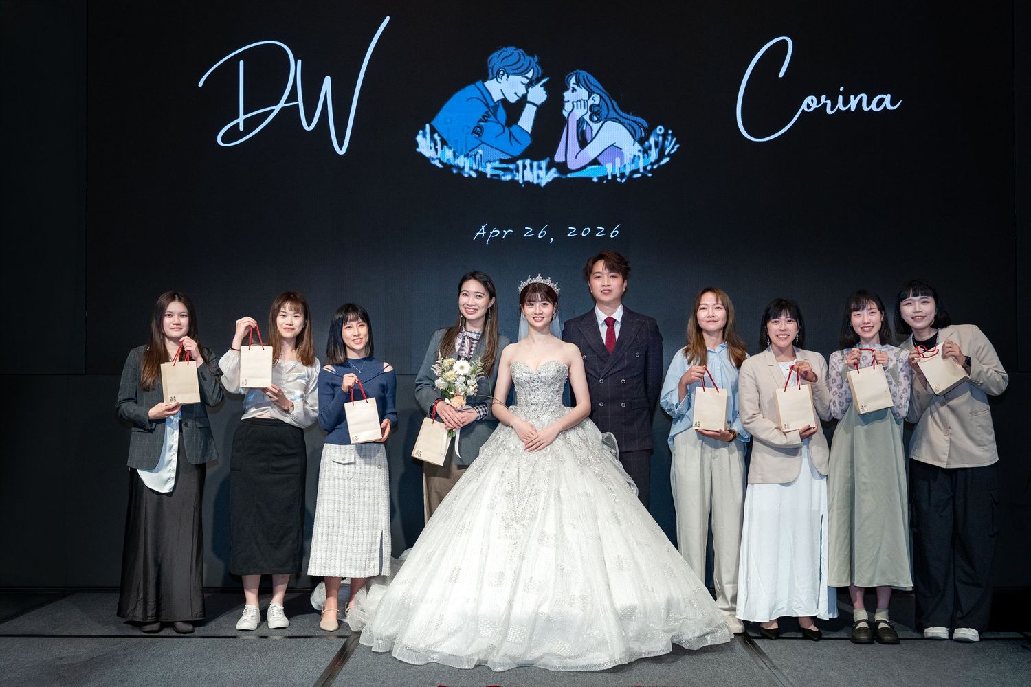 A bride and groom pose with wedding guests at a formal reception, with DW and Corina April 2024 displayed on backdrop.