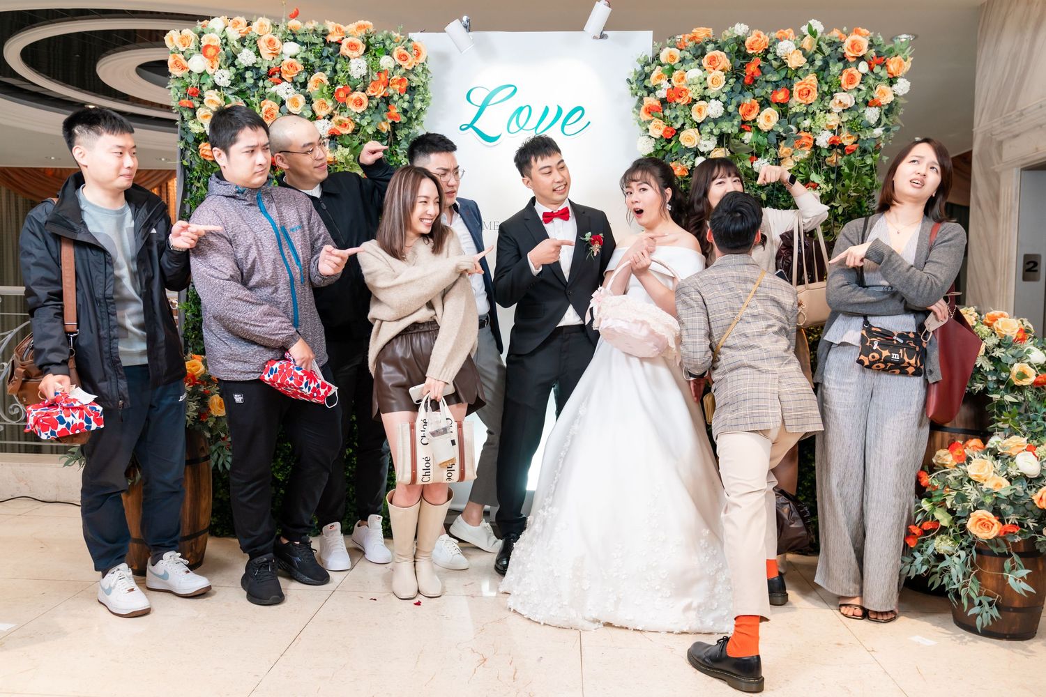 Wedding party poses together in front of floral backdrop with 'Love' sign at reception venue.
