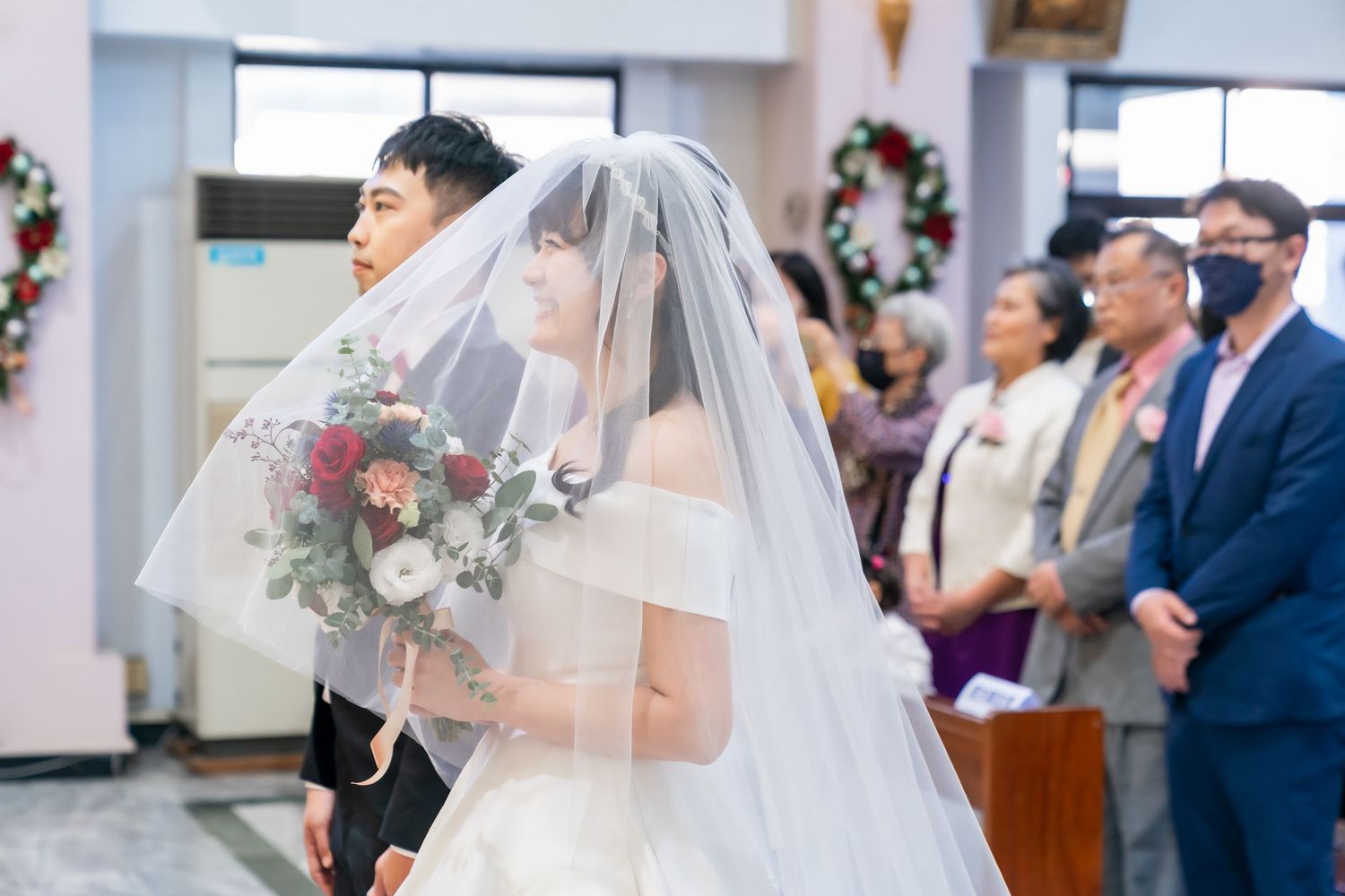 Bride in white dress and veil holding bouquet during church wedding ceremony.