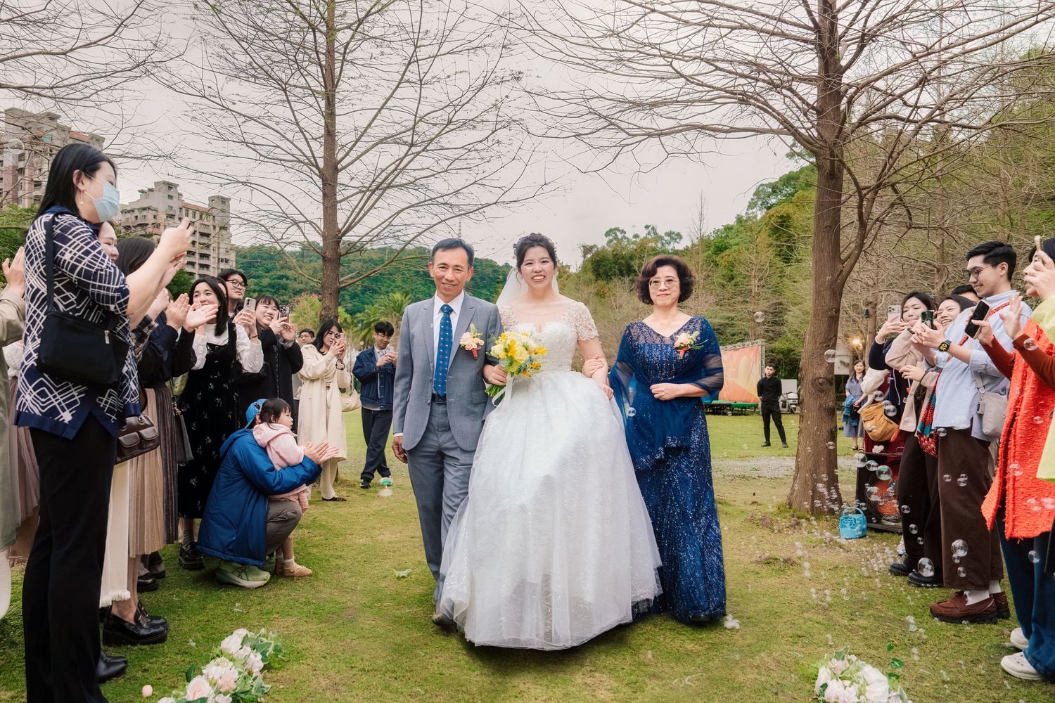 A group of wedding guests gather outdoors under bare trees during a spring celebration.