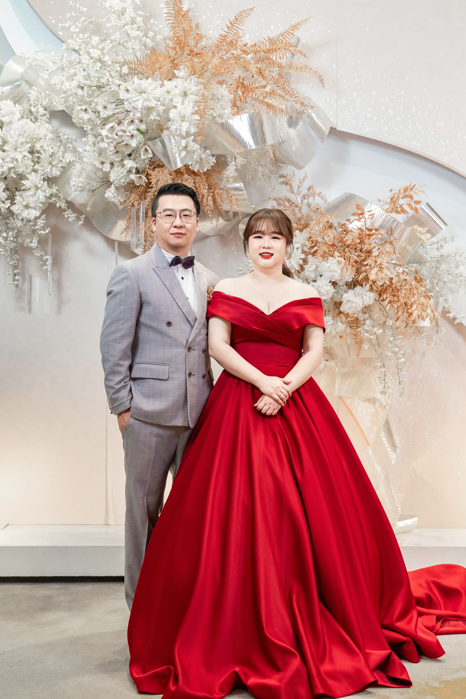 A couple poses in formal attire - a grey suit and red ballgown - in front of a floral arch with dried pampas grass decor.