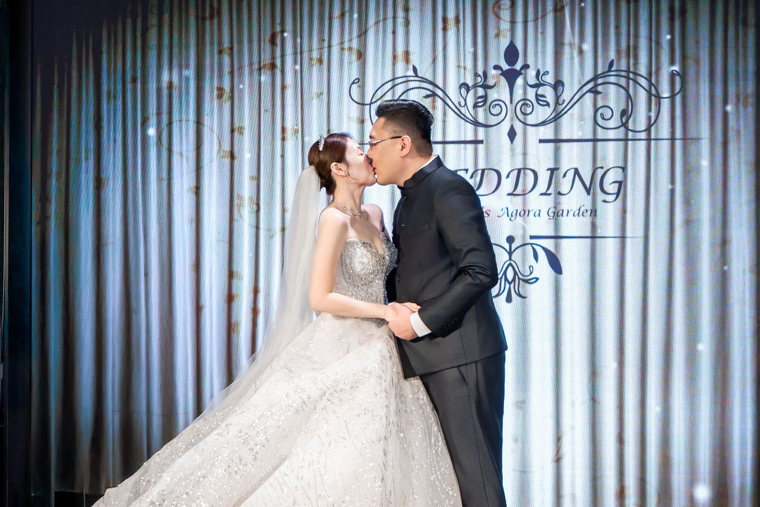 A romantic wedding scene with a couple kissing in front of a blue backdrop with elegant decorative elements.