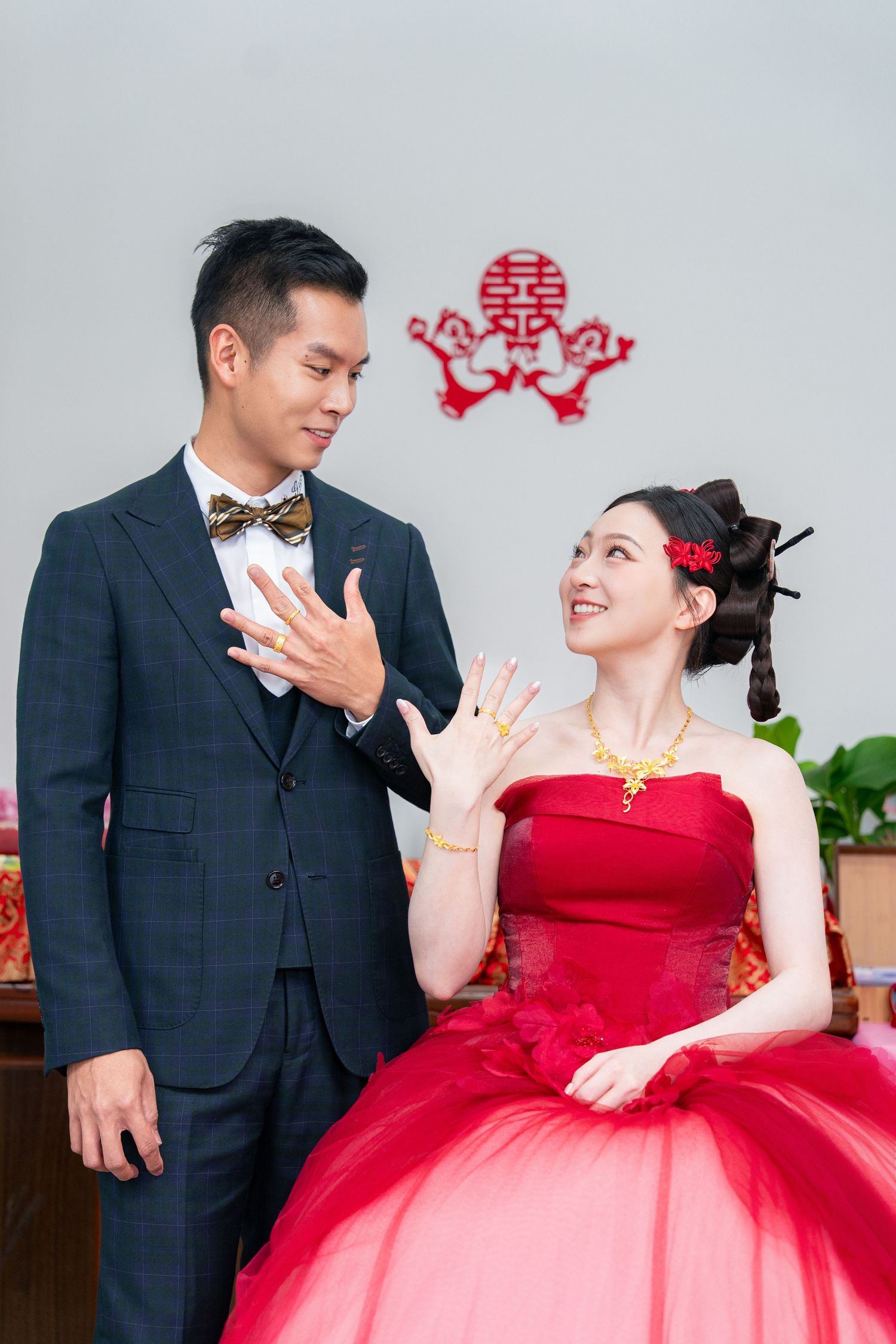Chinese couple in traditional wedding attire displaying rings, bride in red gown with gold jewelry, double happiness symbol behind them.