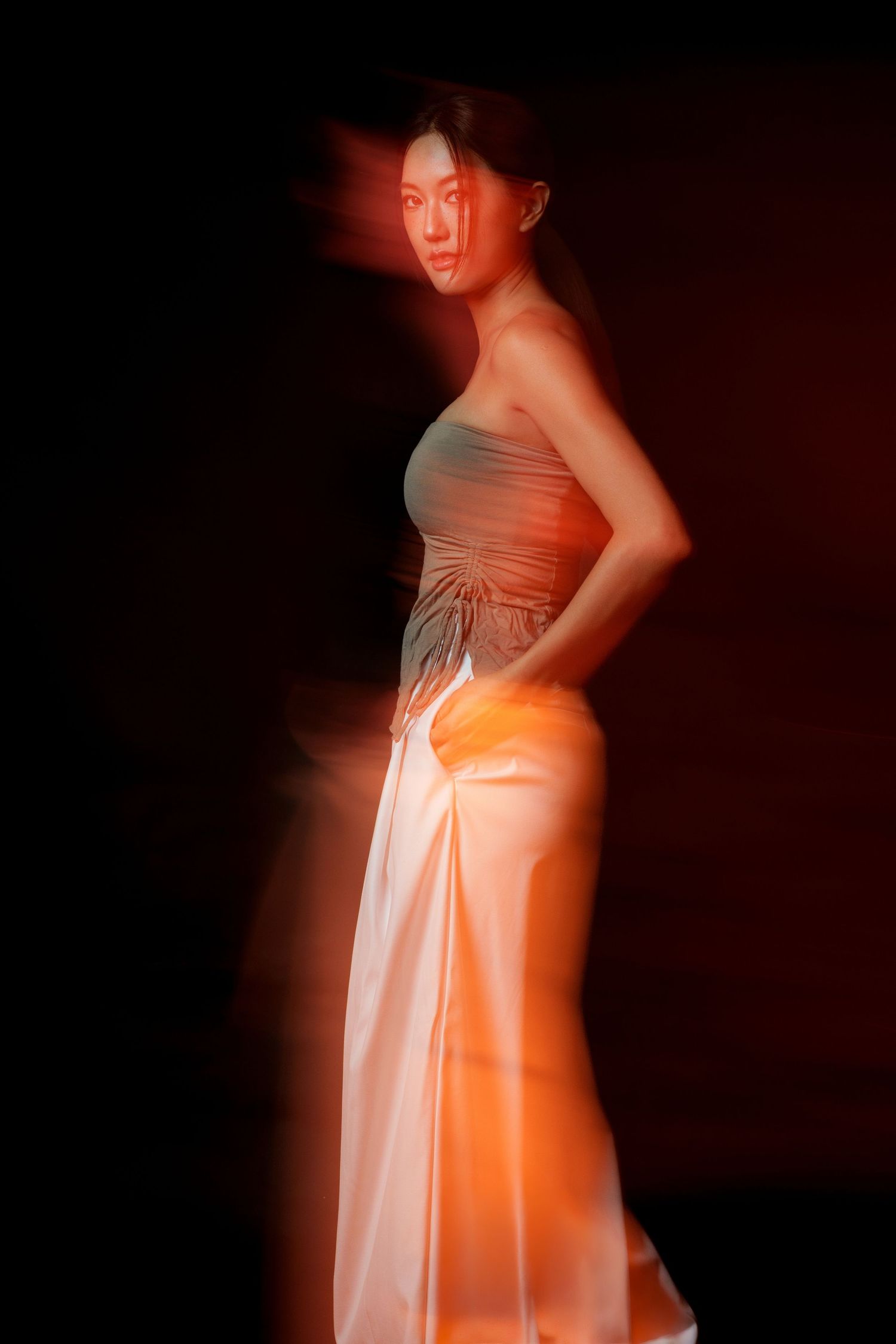 Woman in elegant gray top and white satin skirt poses dramatically under warm orange lighting against dark background.