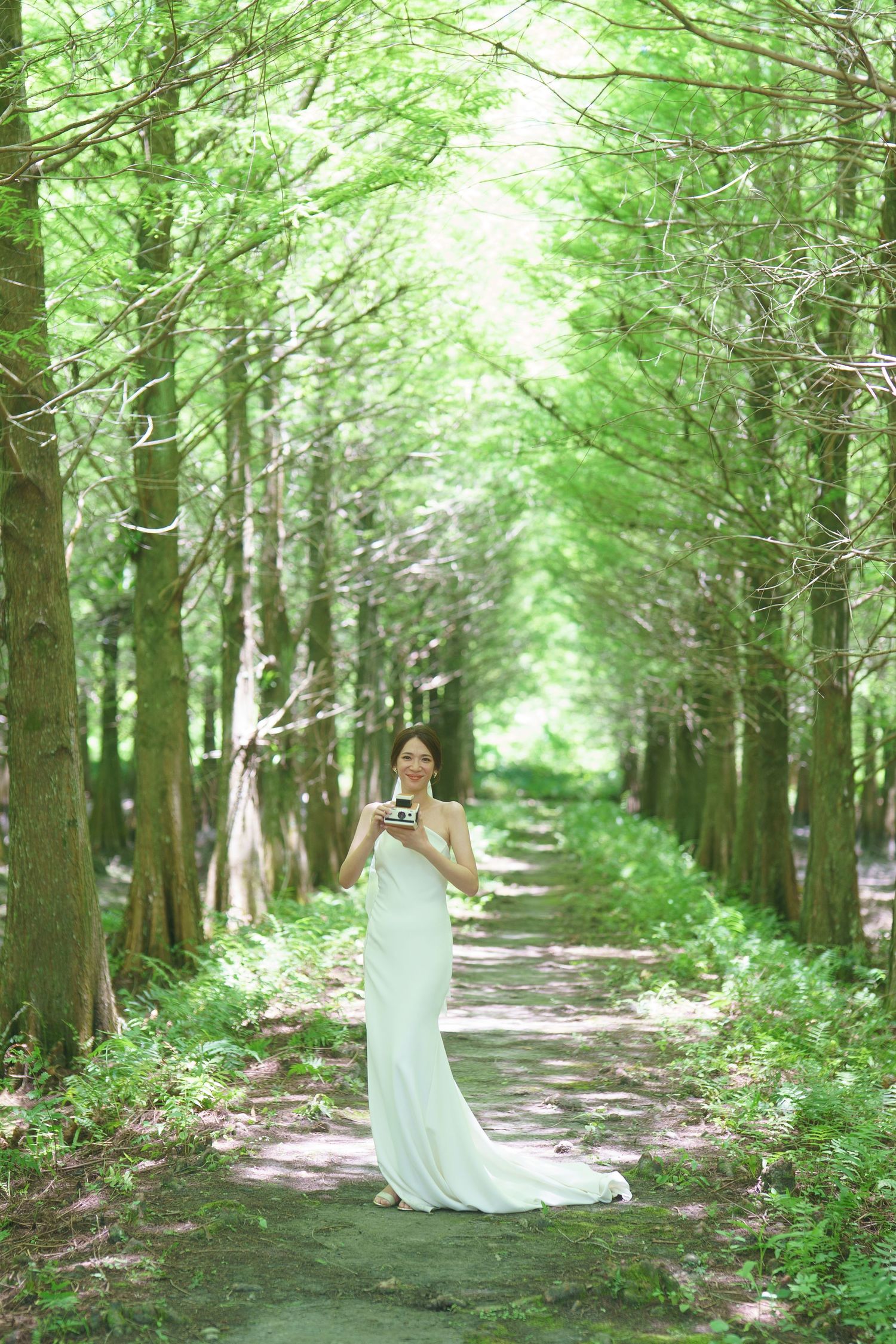 Person in a flowing white gown stands elegantly among tall trees in a sunlit forest path.
