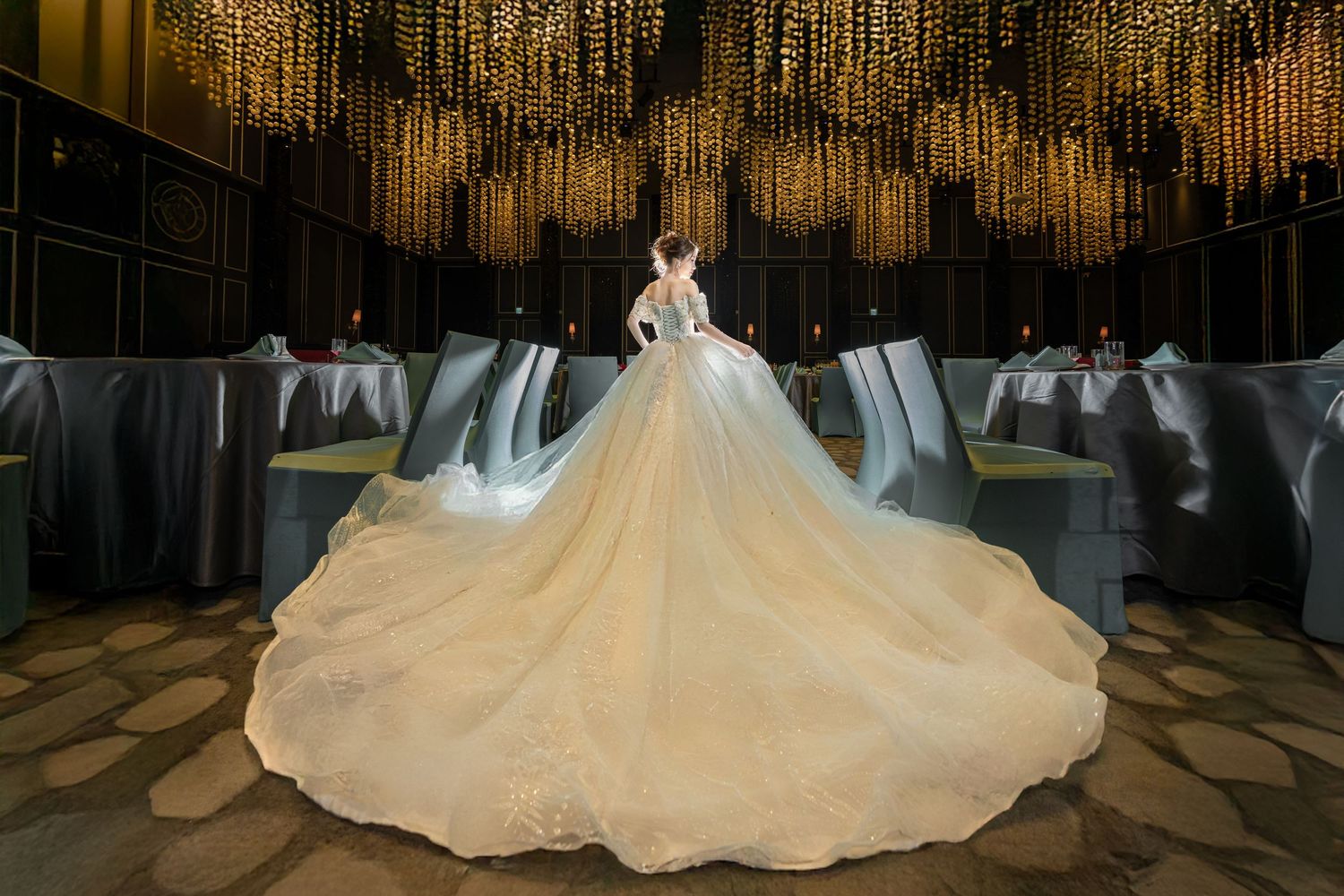 A bride in a flowing white wedding gown stands under sparkling crystal chandeliers in an elegant ballroom.