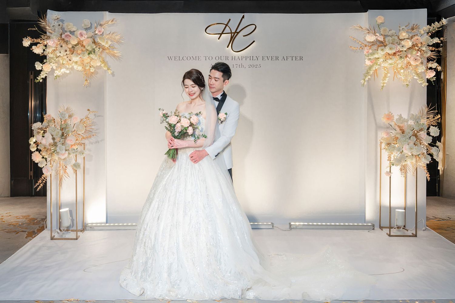 Elegant wedding stage decorated with gold floral stands and lanterns against a white backdrop with illuminated monogram.