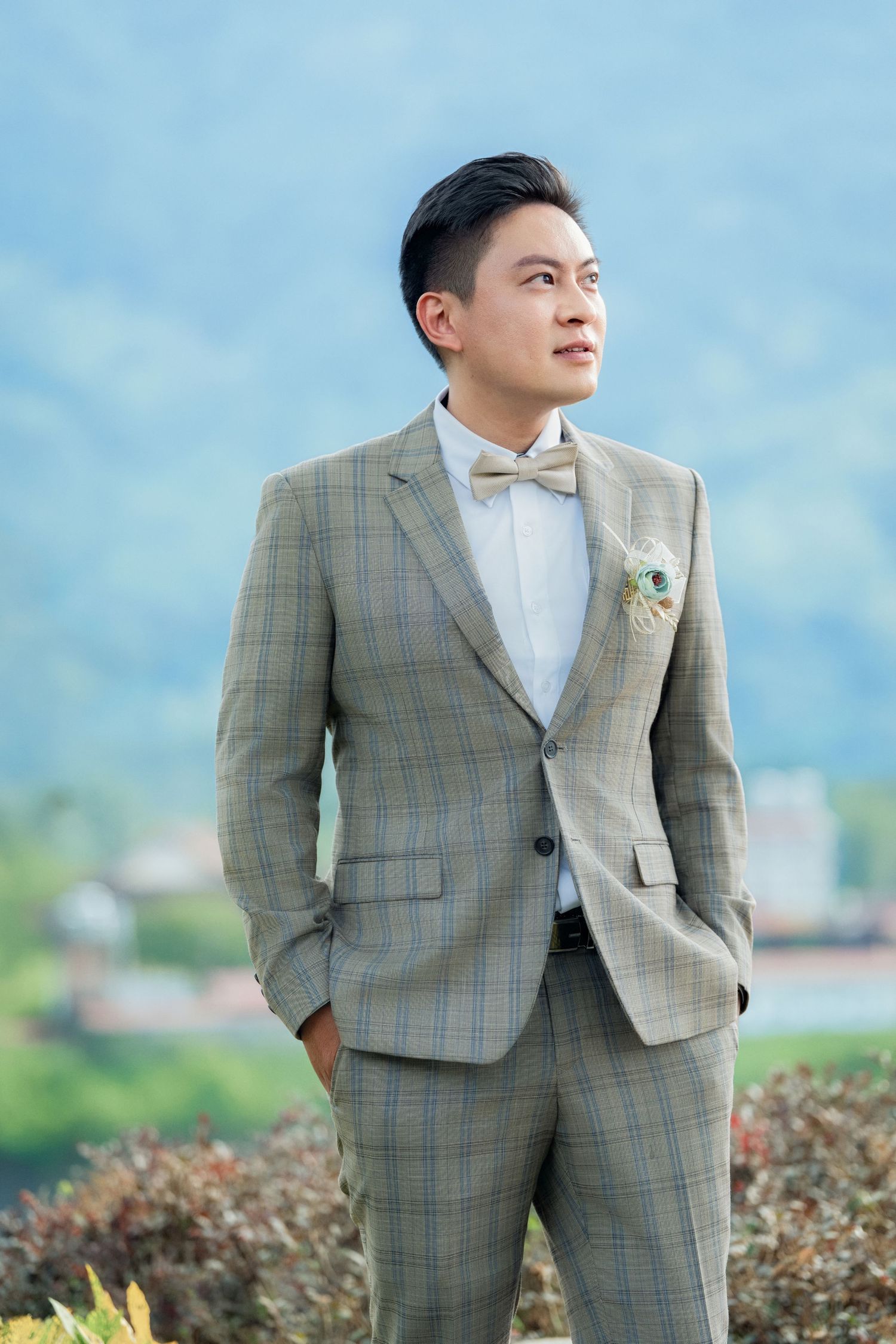A person in a light gray plaid suit and white dress shirt poses in an outdoor setting with greenery and blue sky background.