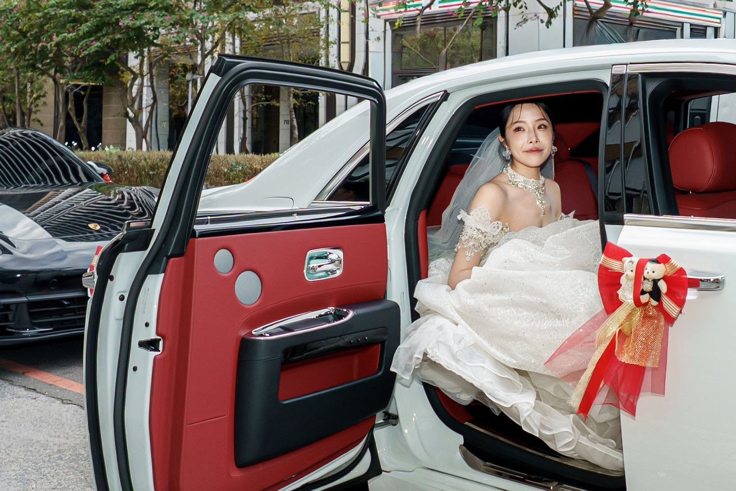 A bride in a white wedding dress sits in a luxury car with red leather interior, holding a red bouquet.