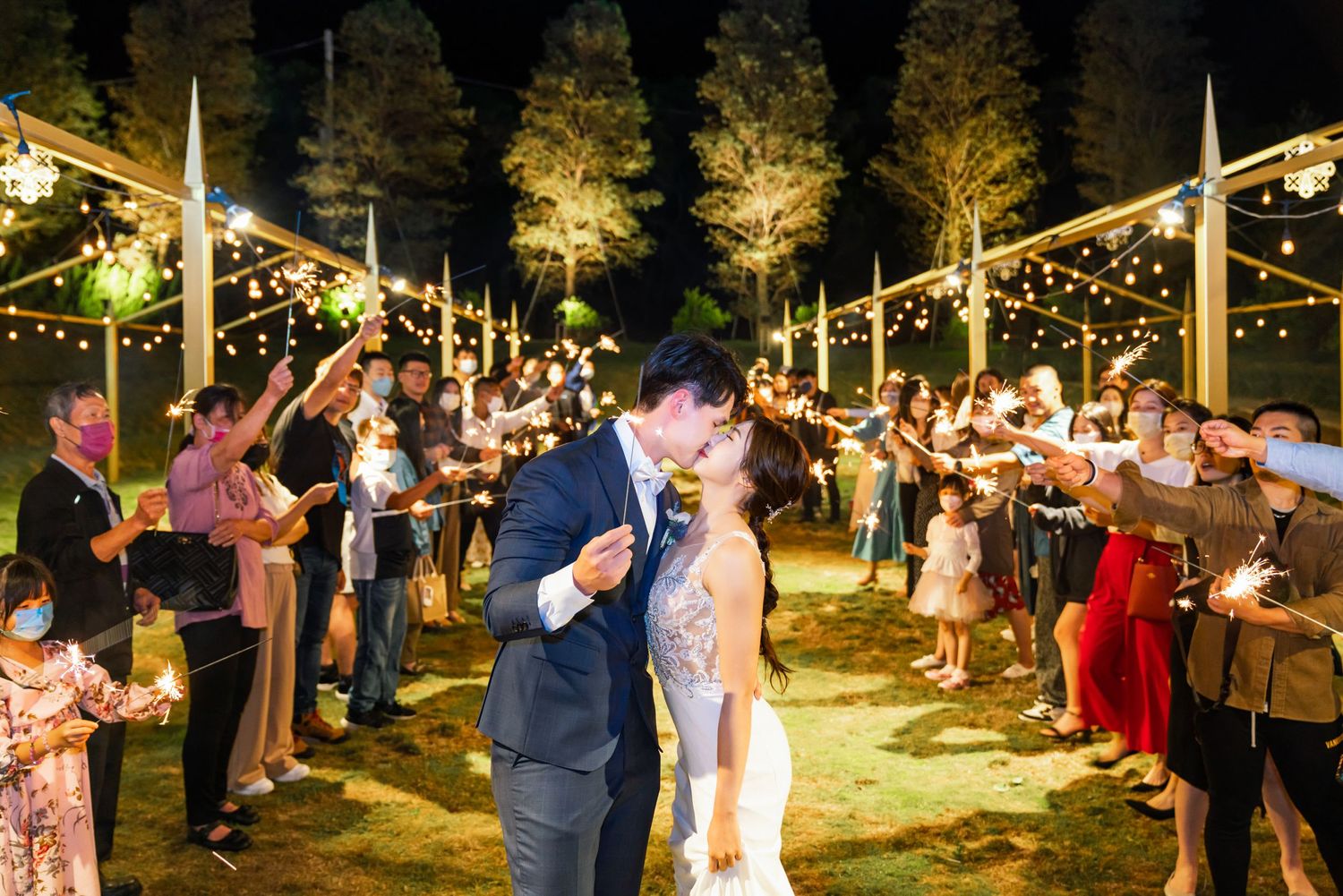 A couple shares their first dance at an outdoor evening wedding reception under romantic string lights with guests looking on.
