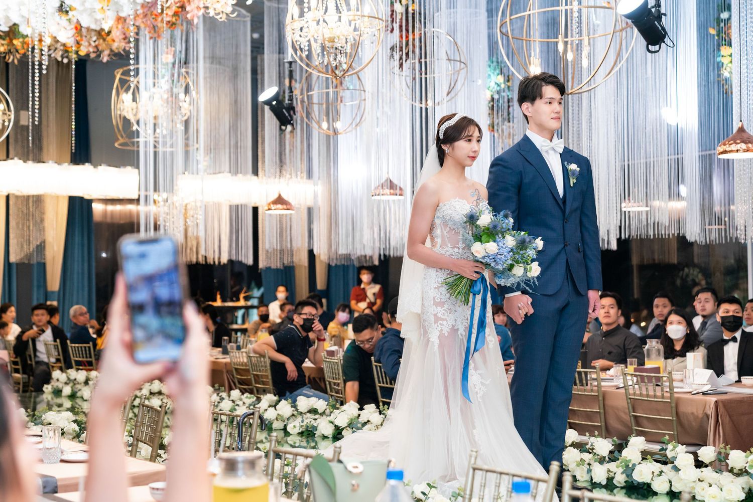 Elegant wedding ceremony in a modern venue with crystal chandeliers, mirrored decor and floral arrangements along the aisle.