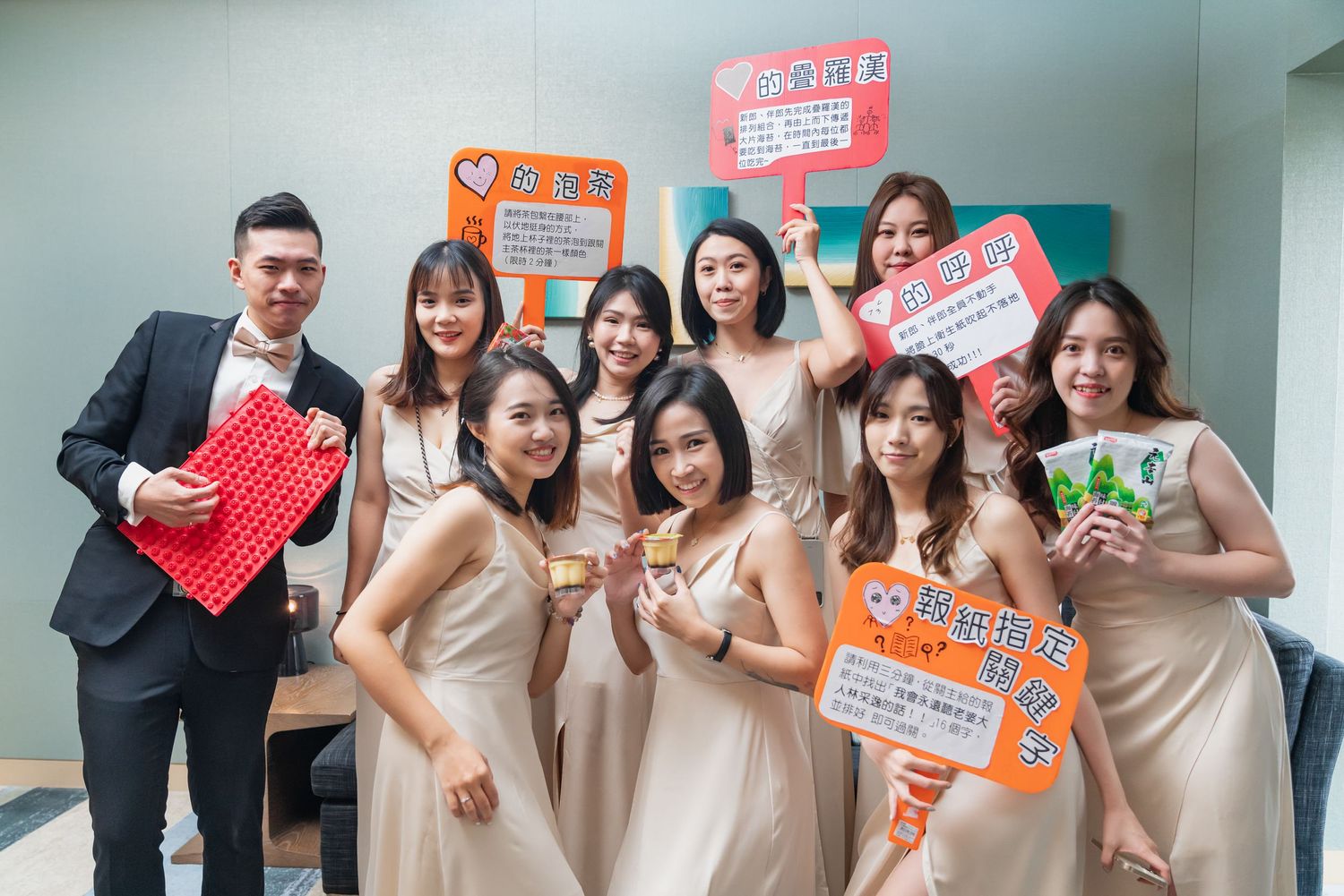 Wedding party members in beige dresses and formal attire pose together while holding colorful signs at an event.