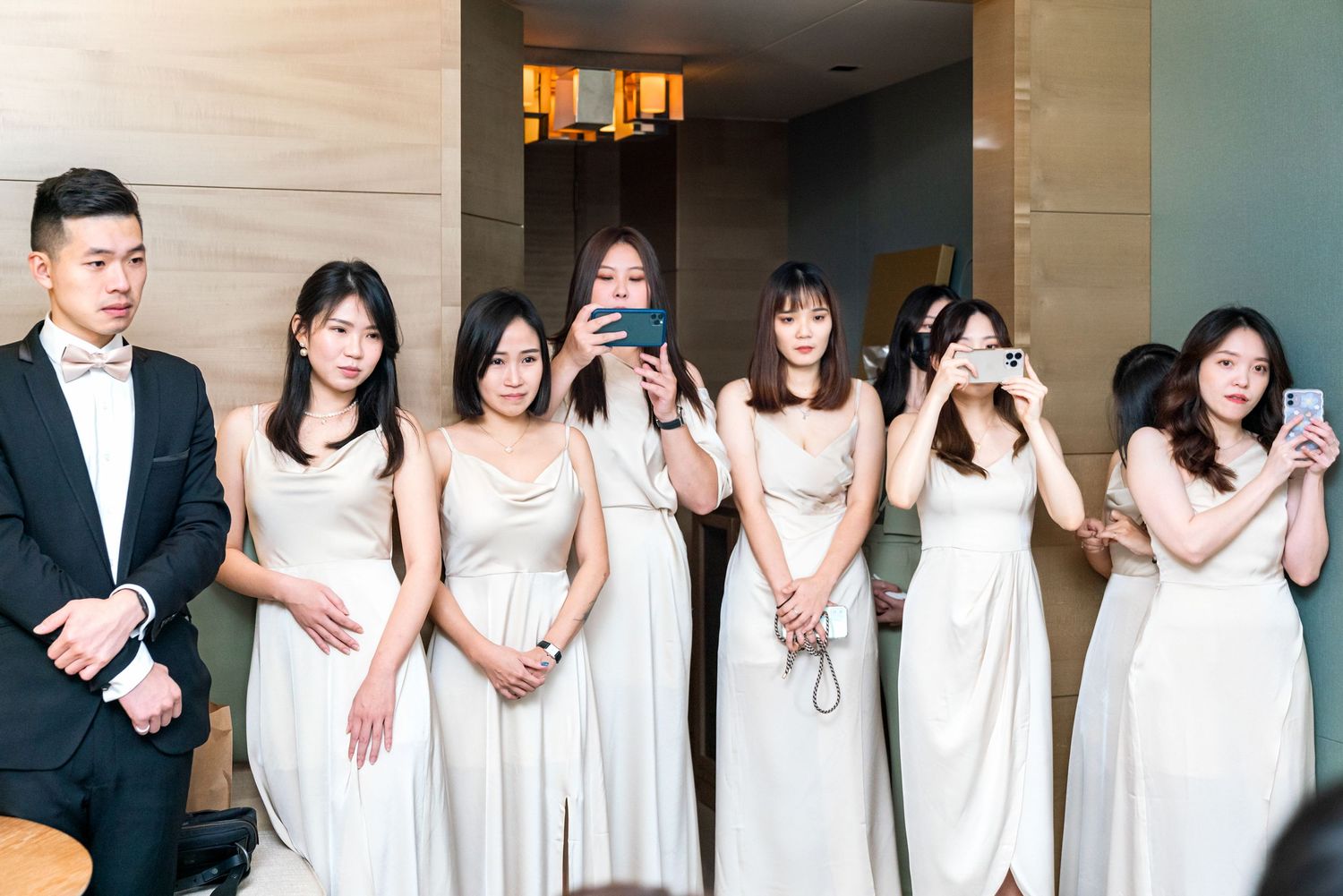 A wedding party lineup in elegant attire with a man in a black suit and others wearing matching white gowns.