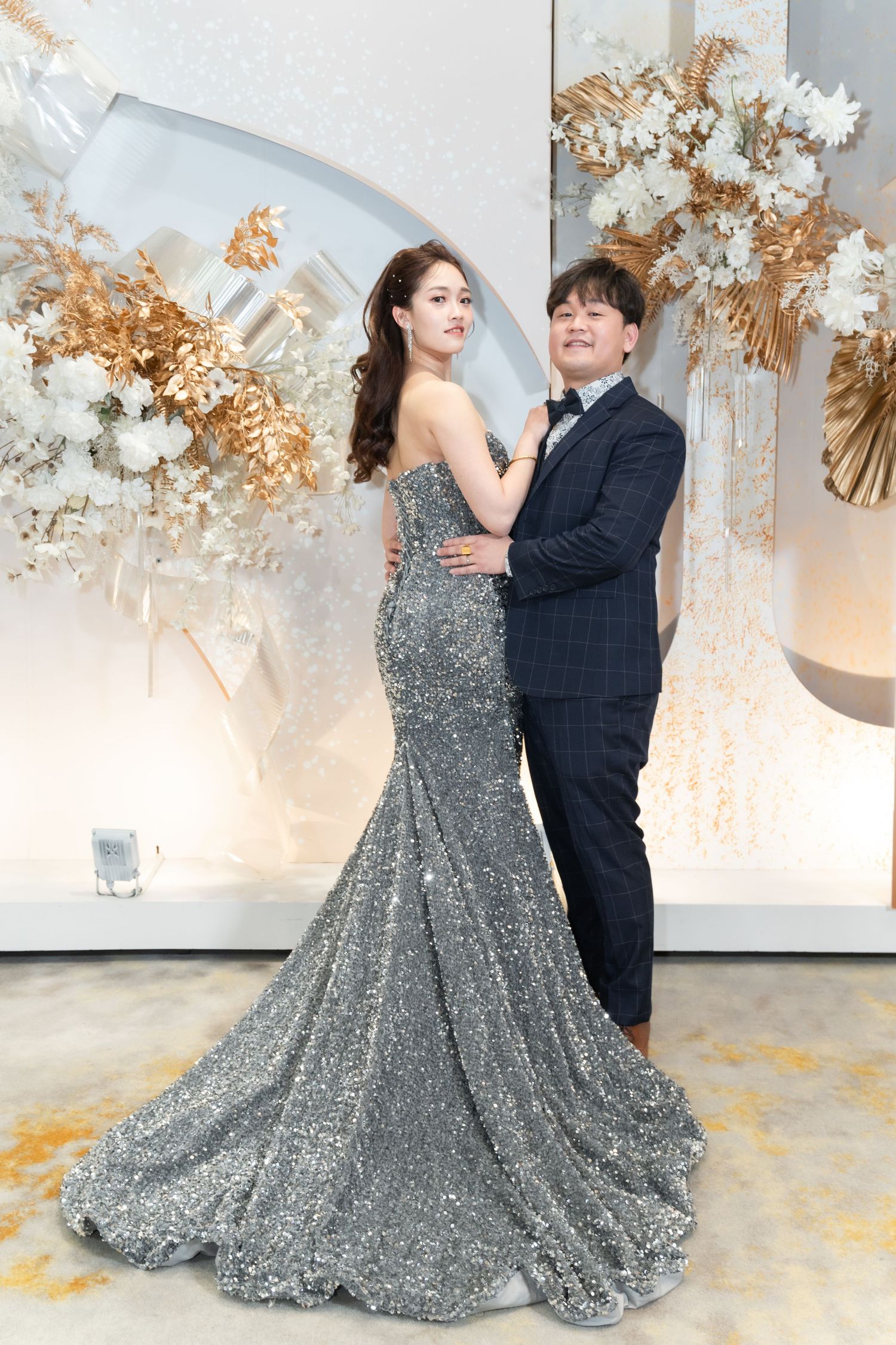 A couple poses together at an elegant event with gold and white floral decorations, the woman wearing a sparkly silver gown.