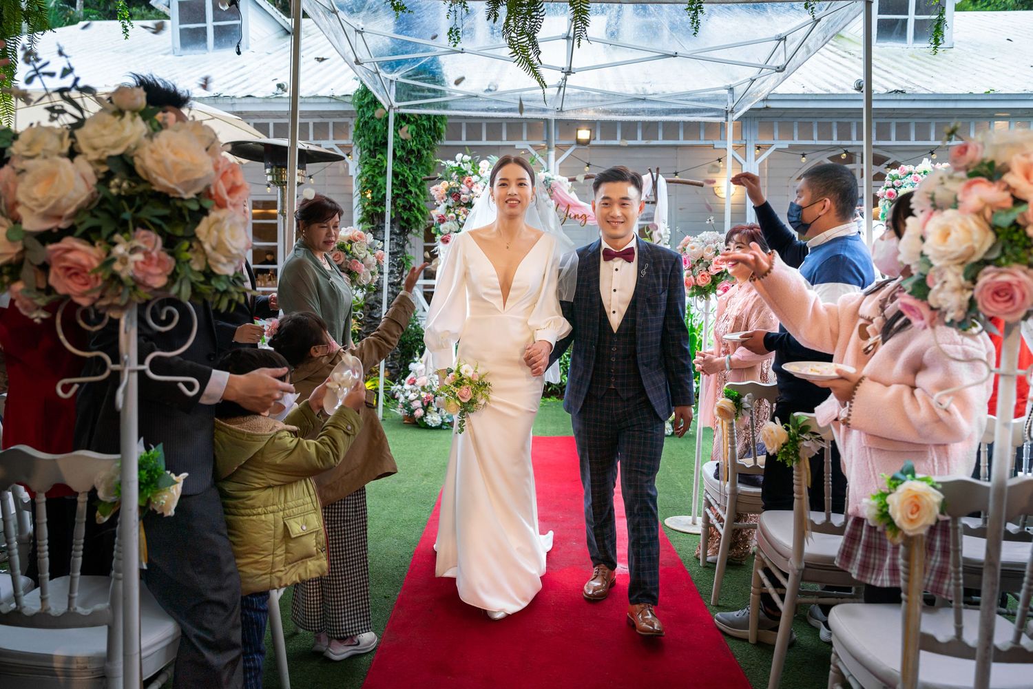 A couple walks down a red carpet aisle runner during an outdoor wedding ceremony with floral decorations and guests.