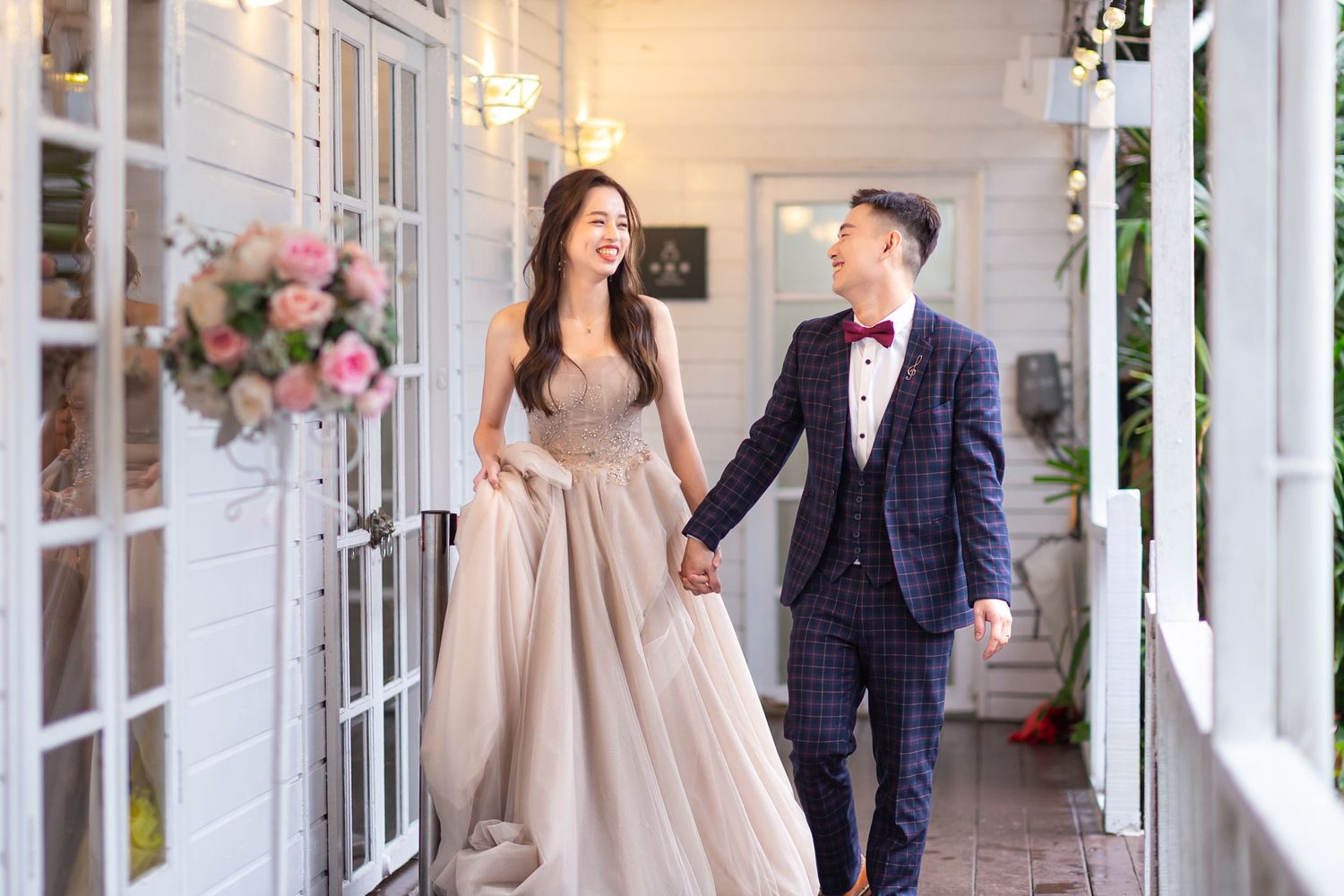 A couple walks hand in hand on a porch decorated with pink flowers, wearing elegant formal attire.
