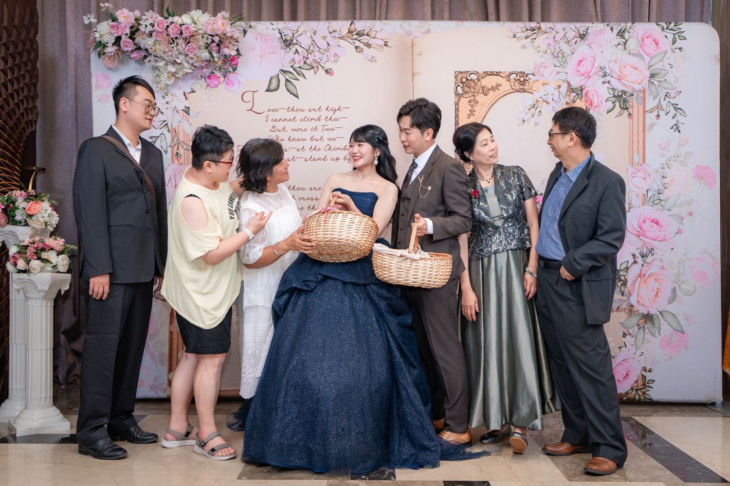 Group of elegantly dressed people gathered at a floral wedding reception backdrop, sharing a joyful moment together.