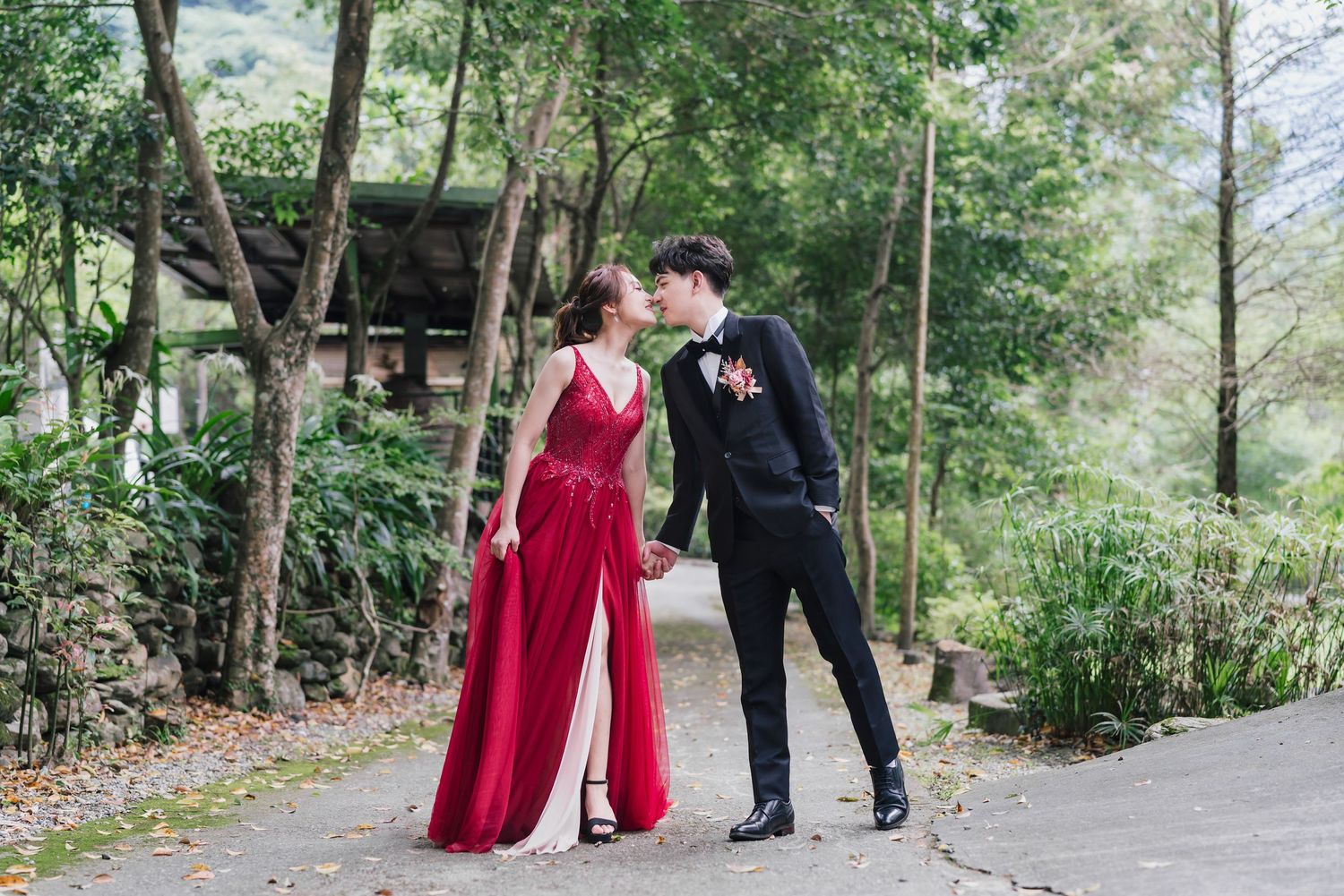 Two figures in formal attire, one in a red gown and one in a black suit, share a tender moment on a garden path.