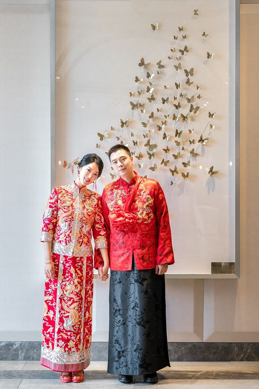 Couple in matching red traditional Chinese wedding attire pose against butterfly wall art.