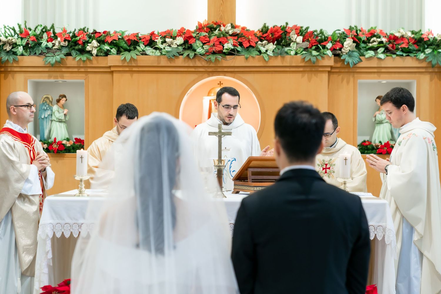Catholic clergy perform wedding ceremony at altar decorated with red and green floral garlands.