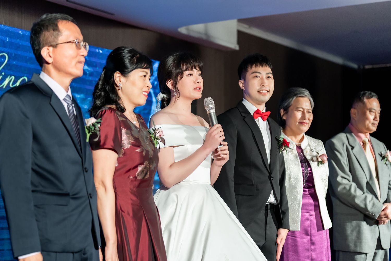 Wedding party lineup on stage with formal attire and blue backdrop.