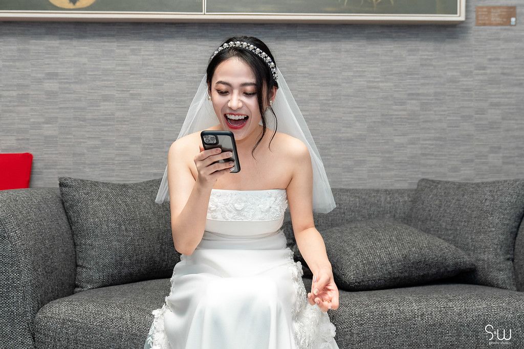 A bride in a white wedding dress sits on a gray couch looking excitedly at her smartphone.