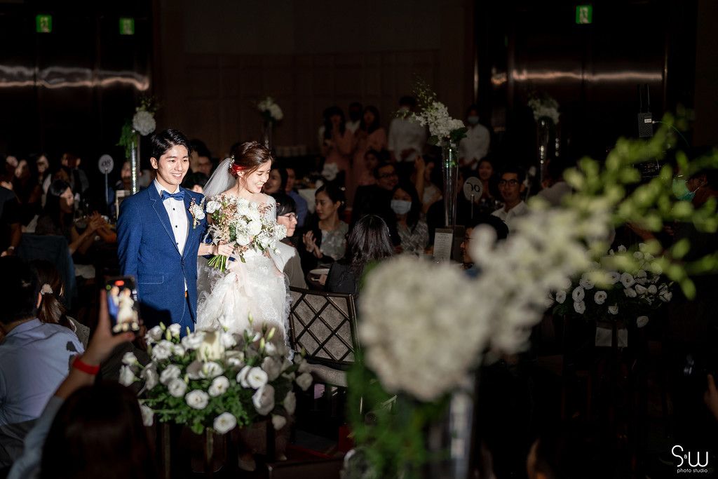 Couple walks during wedding ceremony with elegant white floral arrangements and guests in the background.