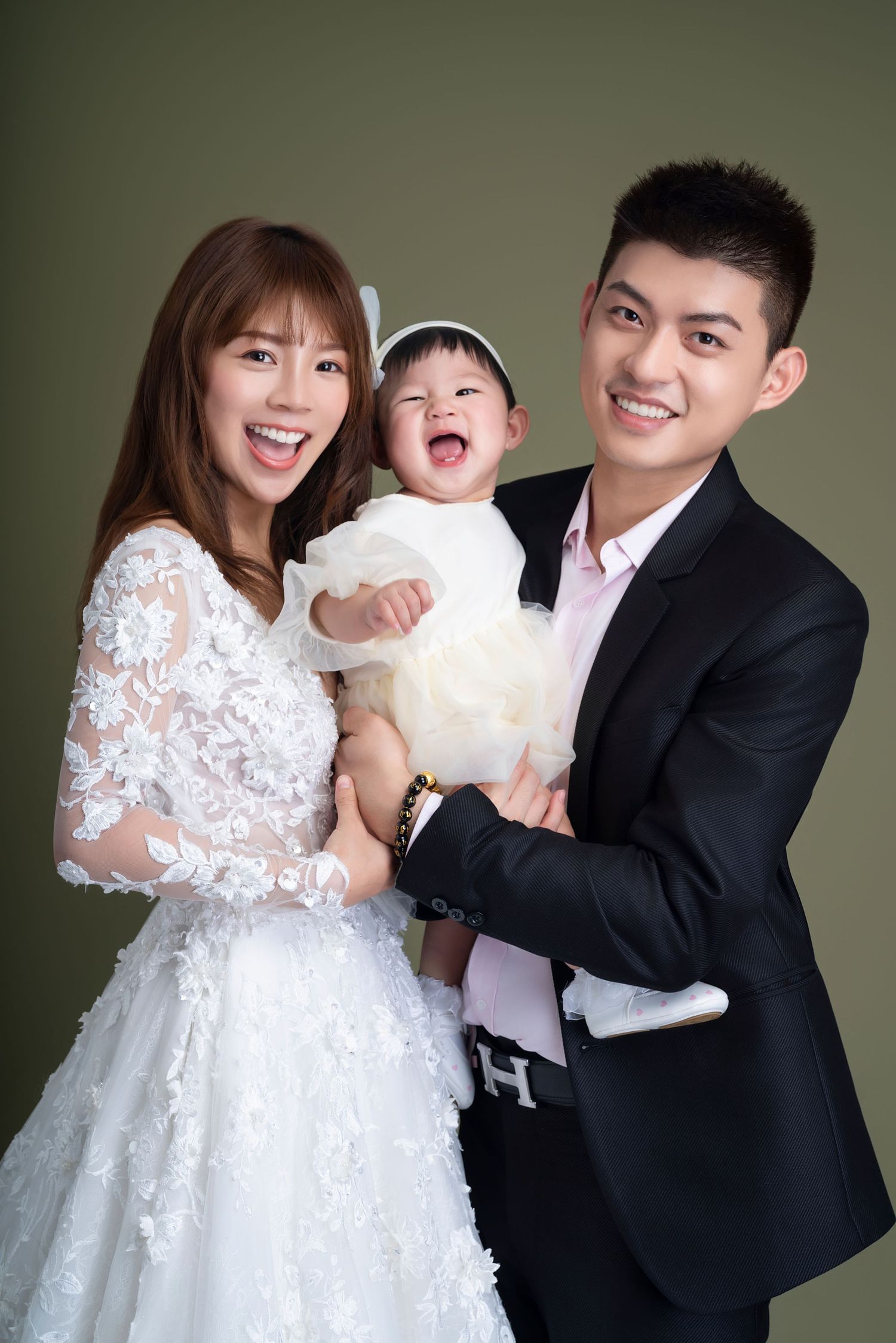 Joyful family portrait featuring a bride in white lace dress, groom in black suit, and laughing baby in formal white outfit.