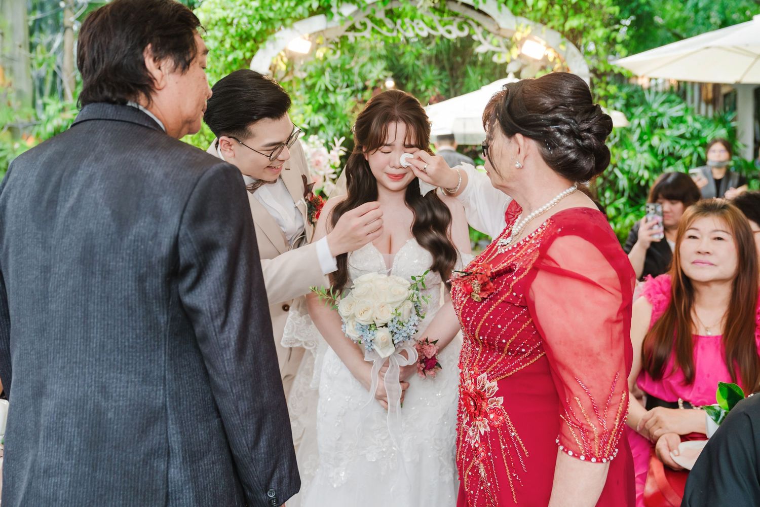 Wedding guests share a joyful moment during an outdoor garden ceremony with lush greenery in the background.