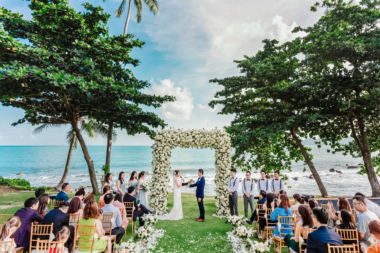 A beachfront wedding ceremony with guests seated under tropical trees and a white floral arch by the ocean.