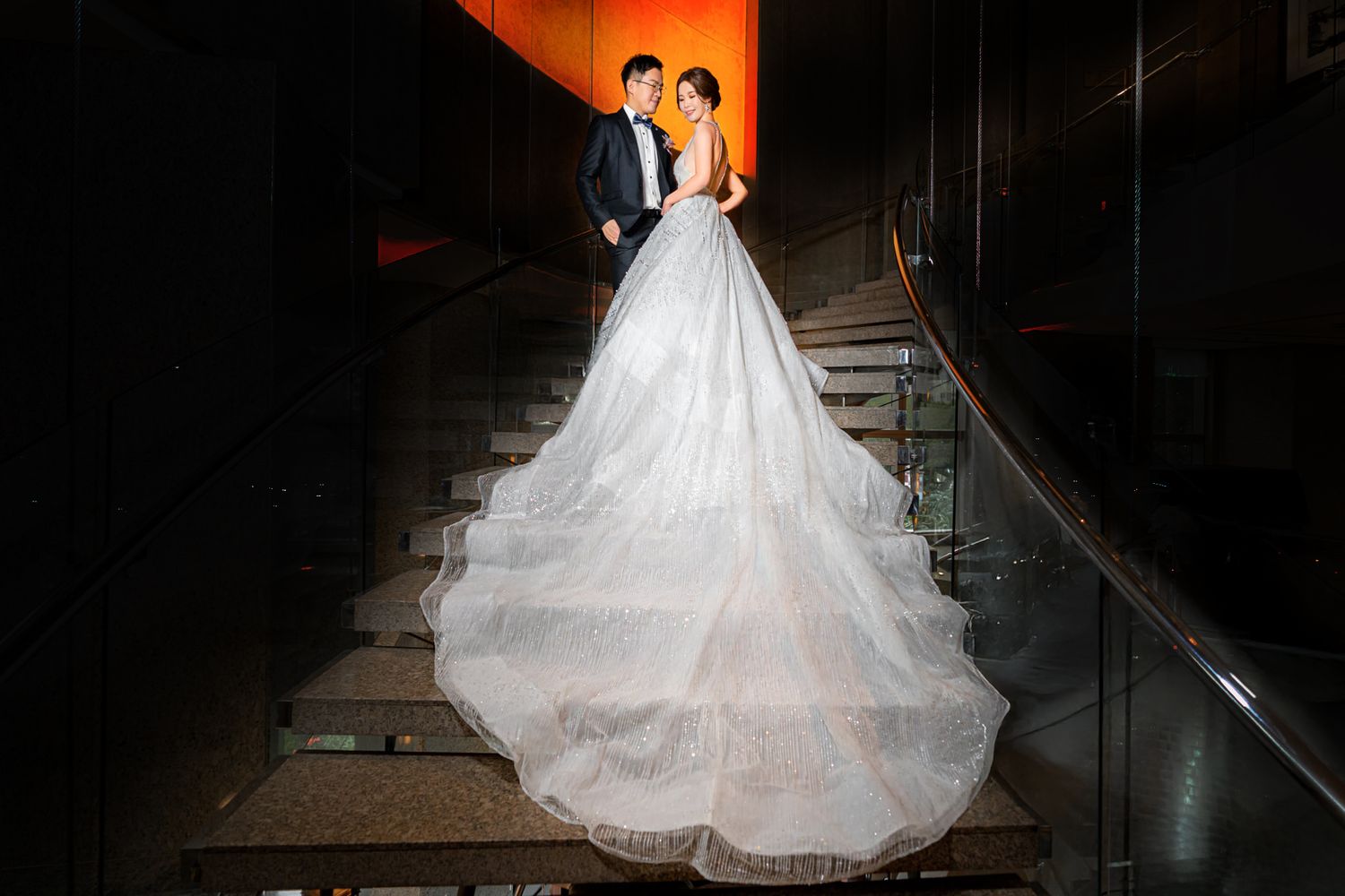 Bride in flowing white ball gown with dramatic long train stands on dark staircase with dramatic orange lighting.