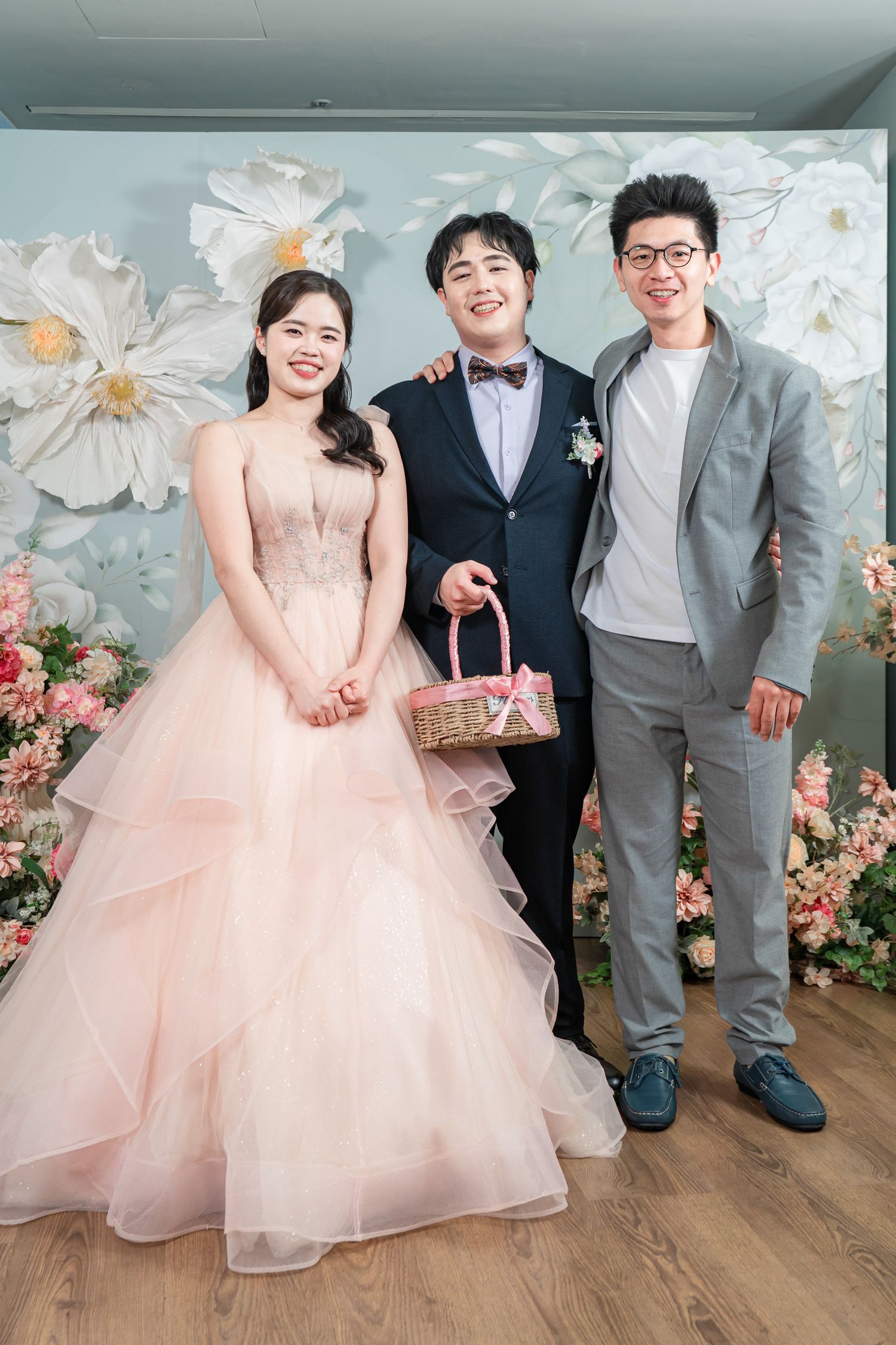 Wedding party poses together in formal attire with bride in pink tulle gown against floral backdrop.