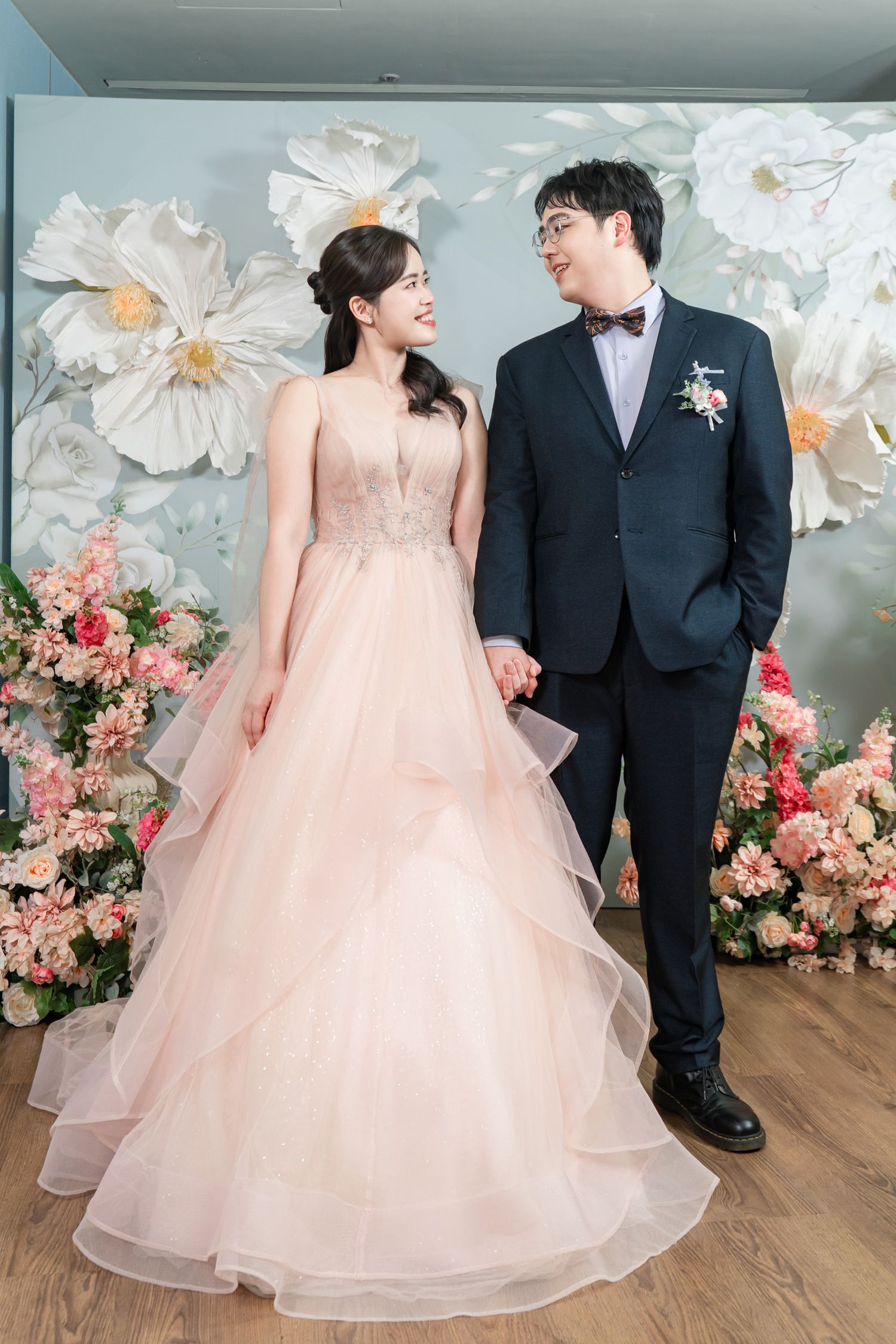 A couple in formal attire stands against a floral backdrop with white and pink blooms for wedding photos.