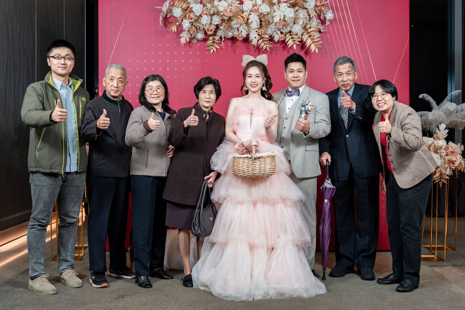 Wedding party members pose together against a pink wall at a celebration venue.