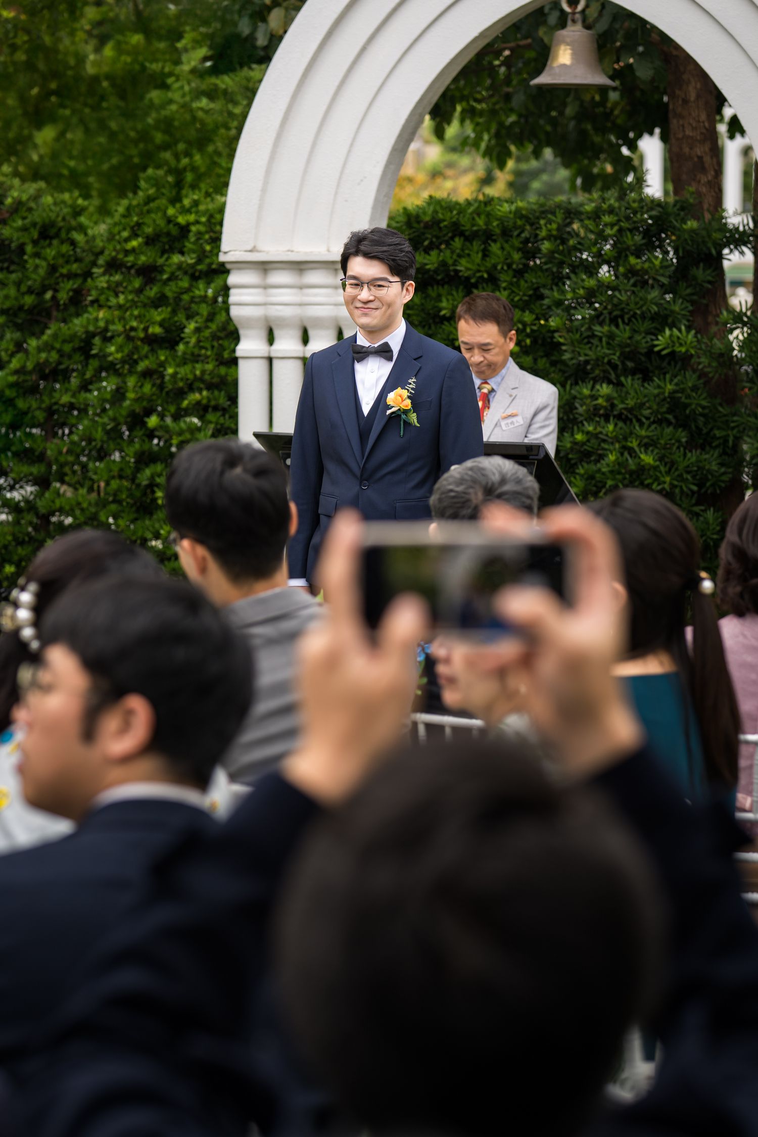 Wedding guests take photos with phones at an outdoor ceremony under a white archway with greenery.