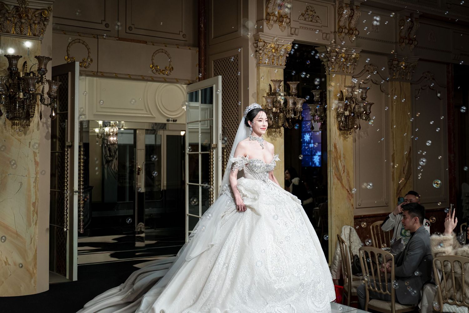 A bride in an elegant white ball gown poses in an ornate room with gilded details and crystal chandeliers.
