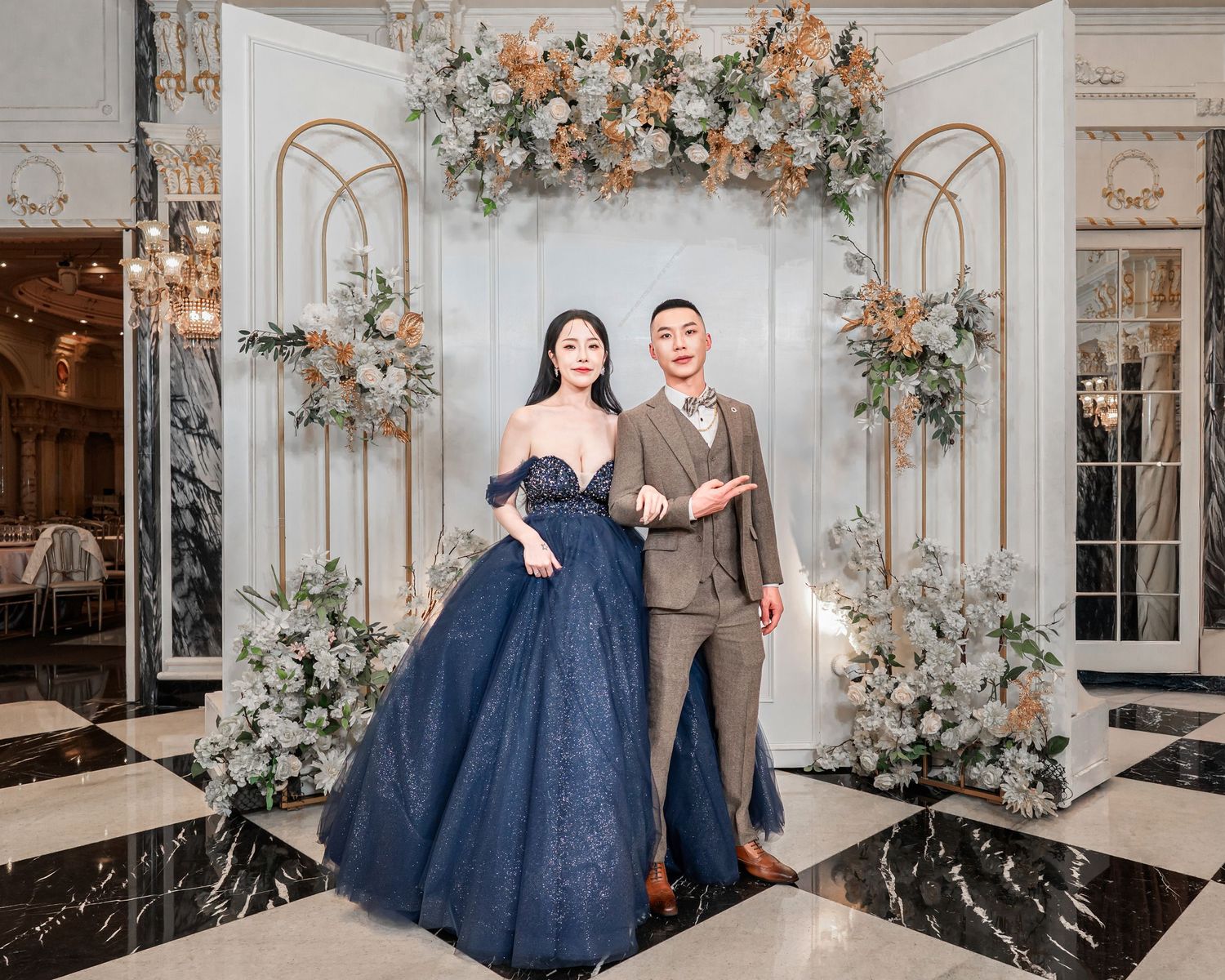 A couple poses in a luxurious wedding venue decorated with elegant floral arrangements and gold arches.