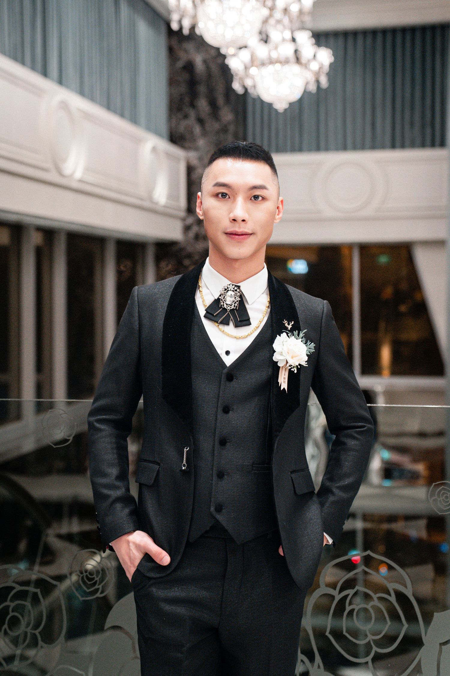 A person wearing a black formal tuxedo suit stands in an elegant venue with crystal chandeliers in the background.