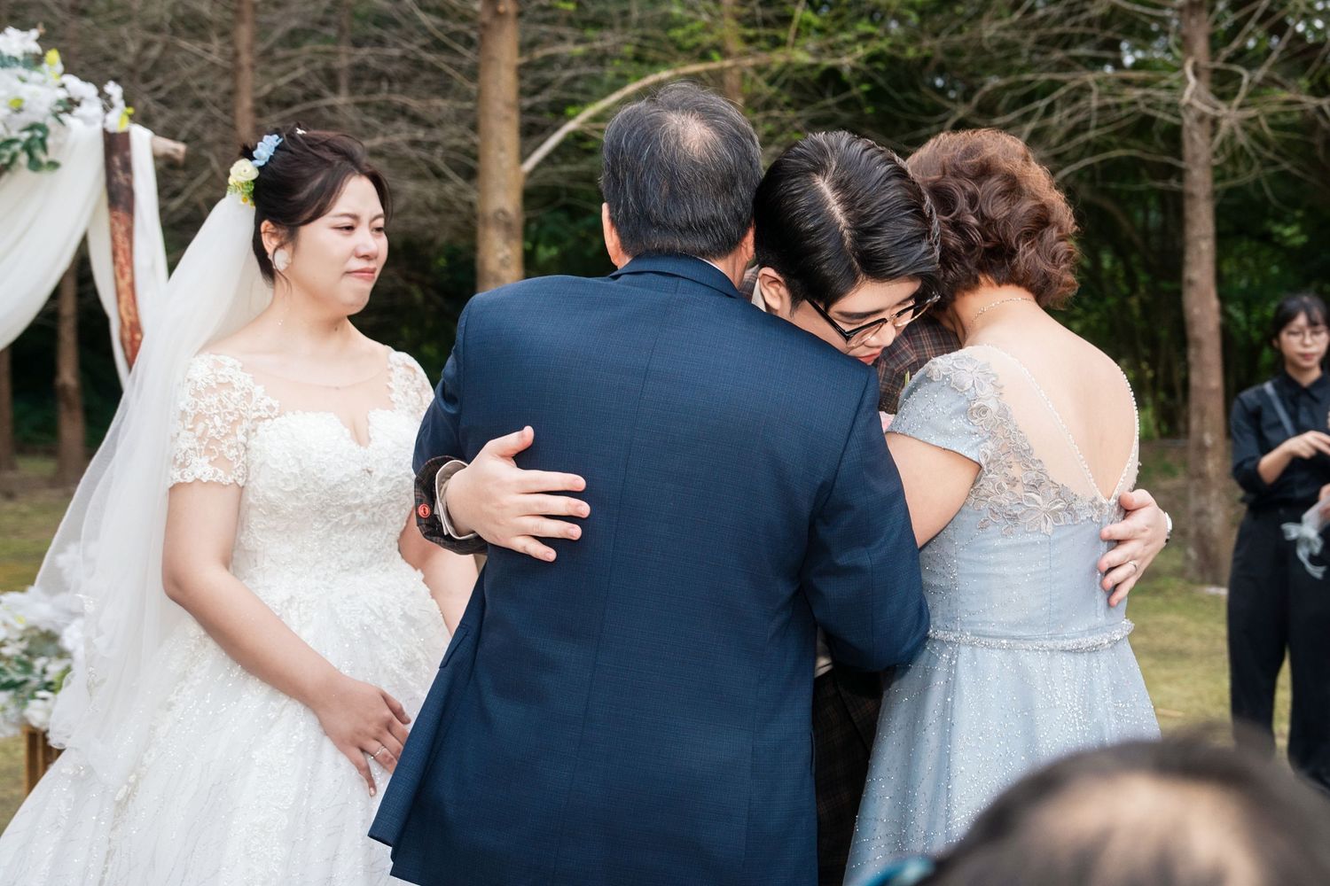 A wedding party gathers outdoors for emotional embraces among trees and natural scenery.