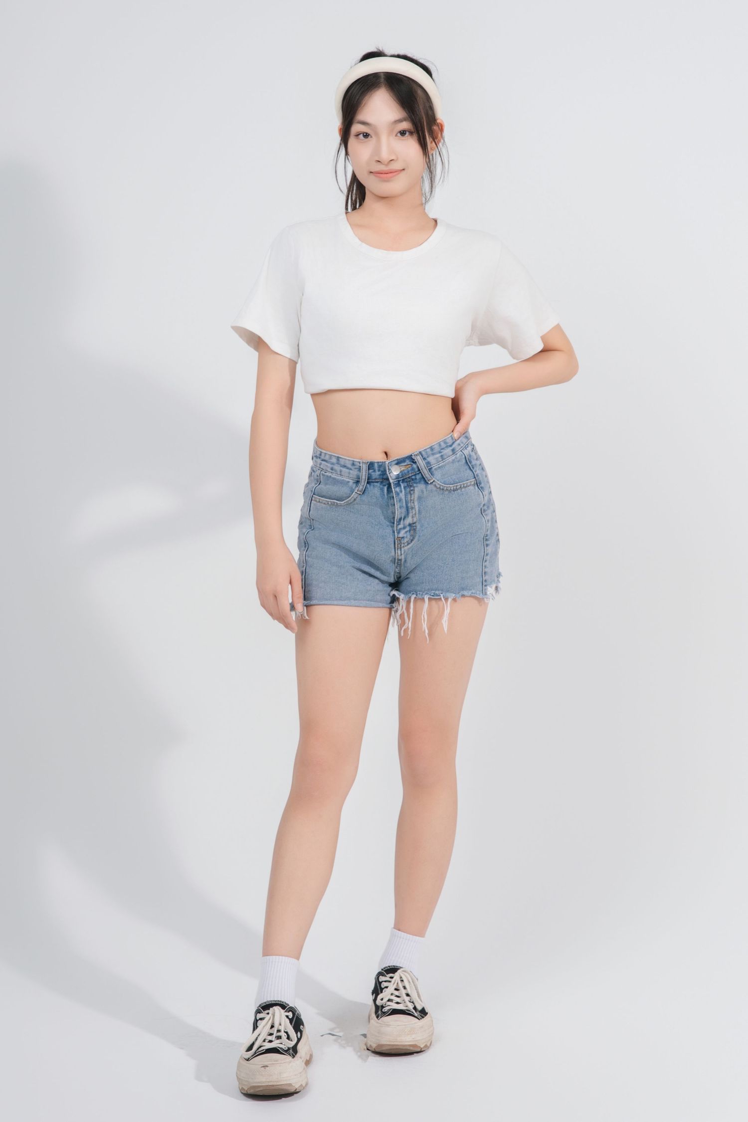 Model in white crop top and distressed denim shorts poses against white background in casual summer outfit.
