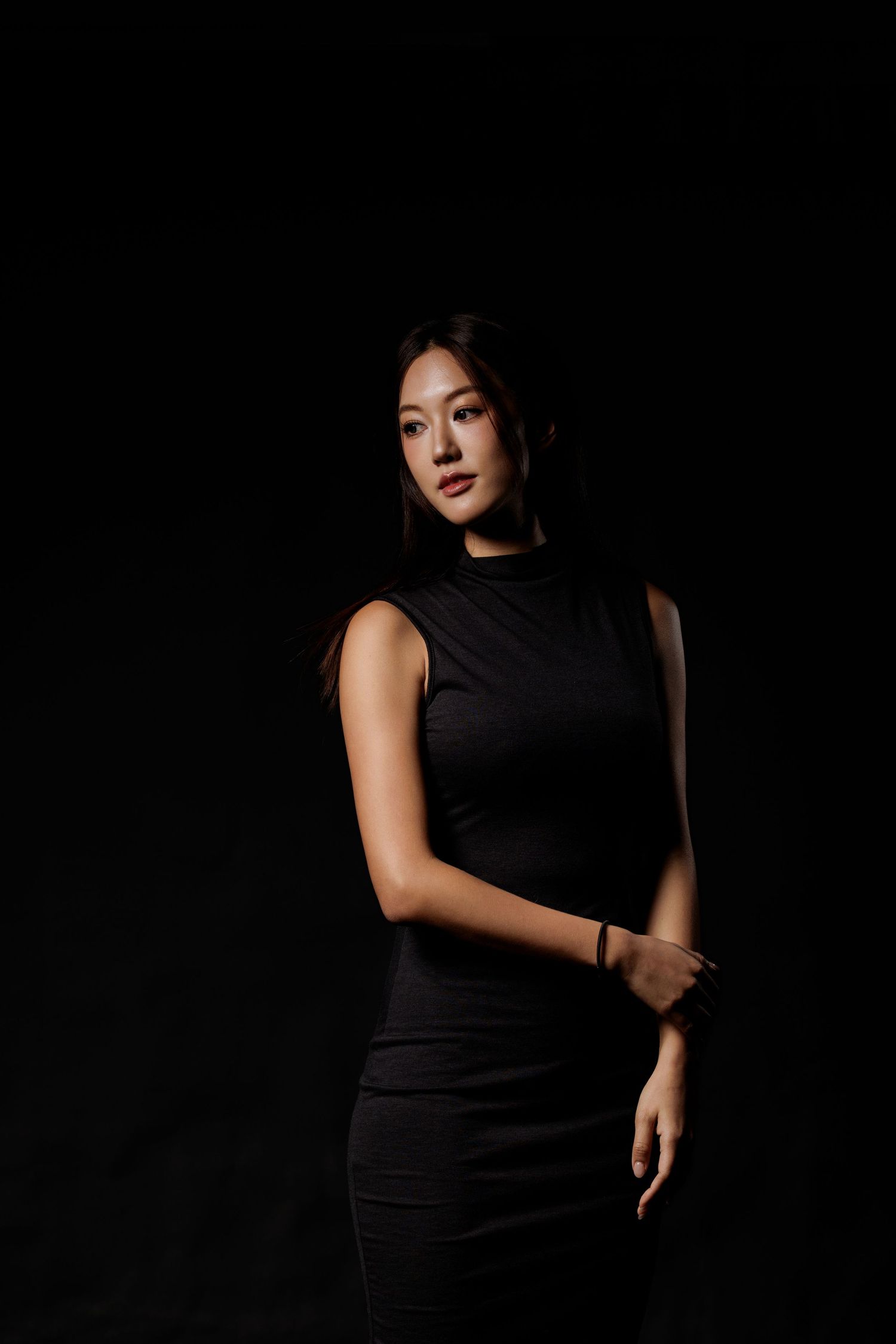 Young Asian woman in sleek black sleeveless dress poses confidently against dark studio background.