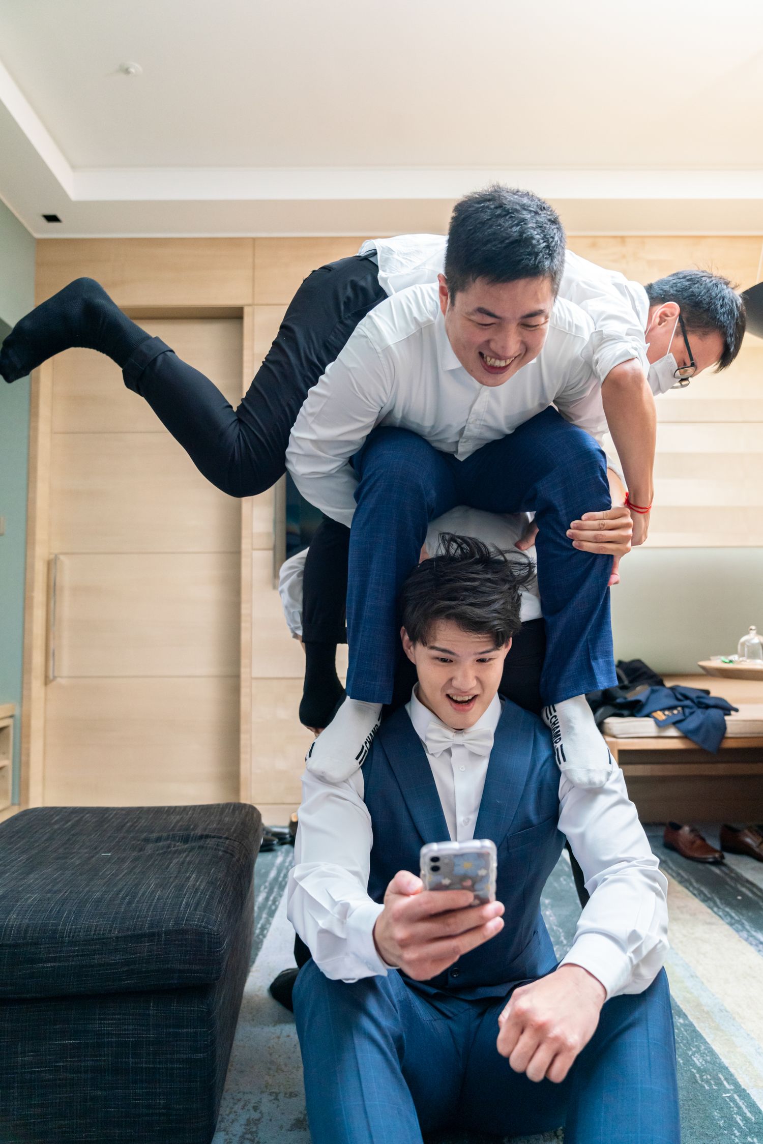 People in suits and casual wear playfully stacking on top of each other while looking at a phone in a living room setting.