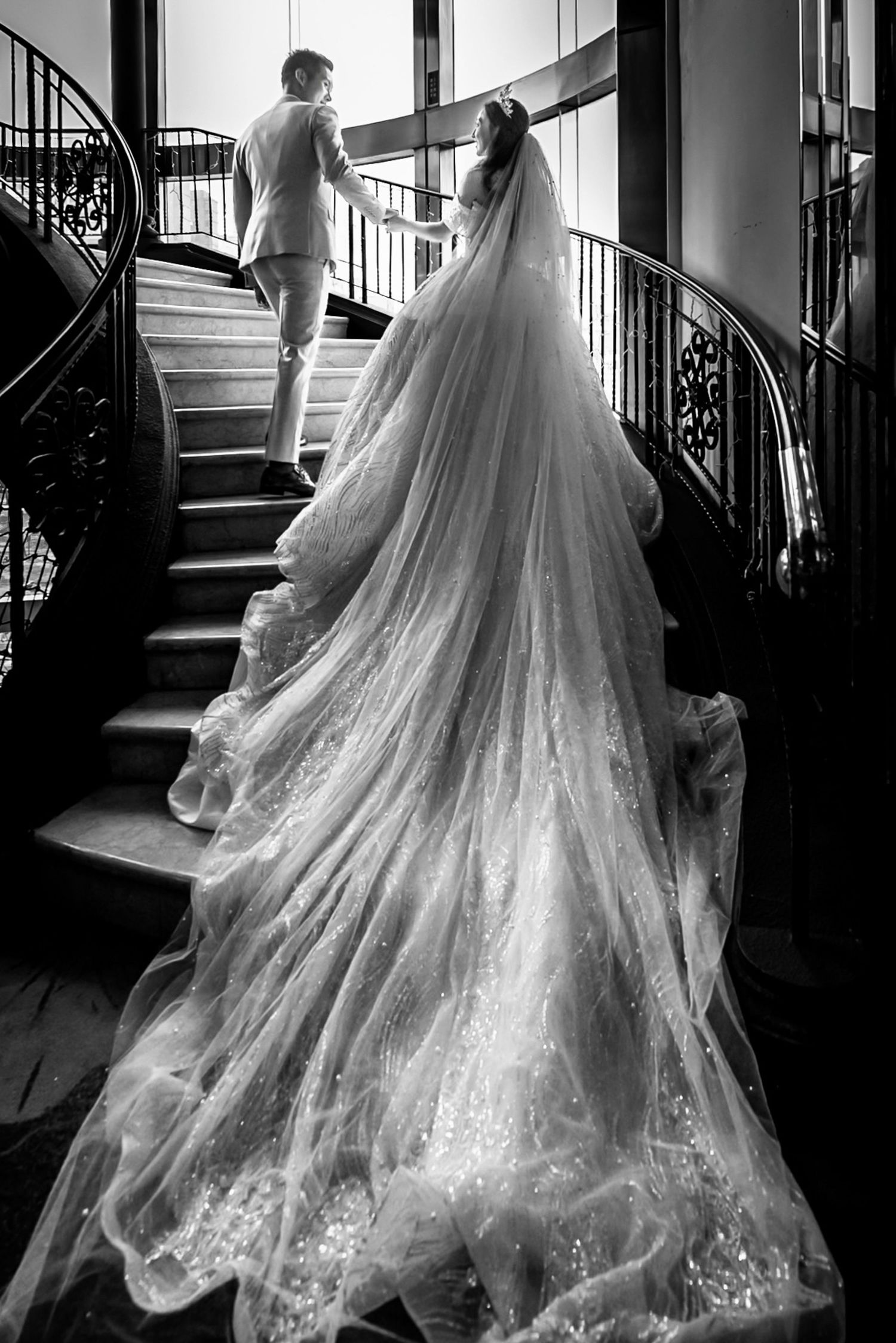 Black and white photo of bride in flowing gown with long veil ascending spiral staircase with groom.