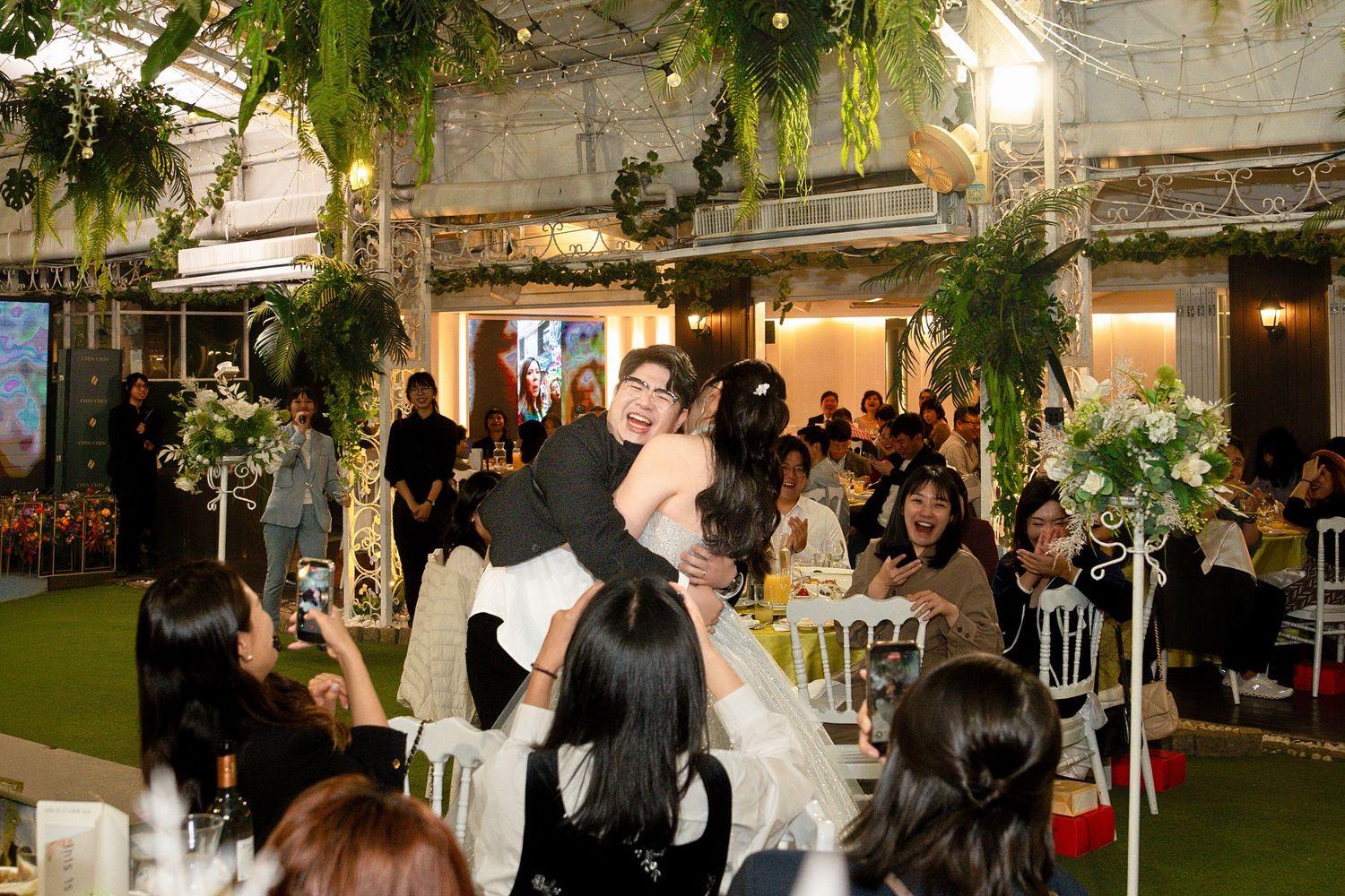 A wedding reception celebration with guests dancing and mingling under hanging greenery and string lights in an indoor venue.
