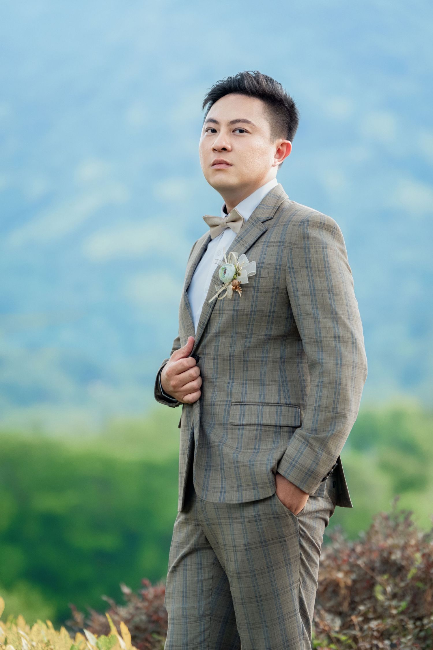 A person wearing a plaid gray suit poses outdoors against a scenic mountain landscape on a sunny day.