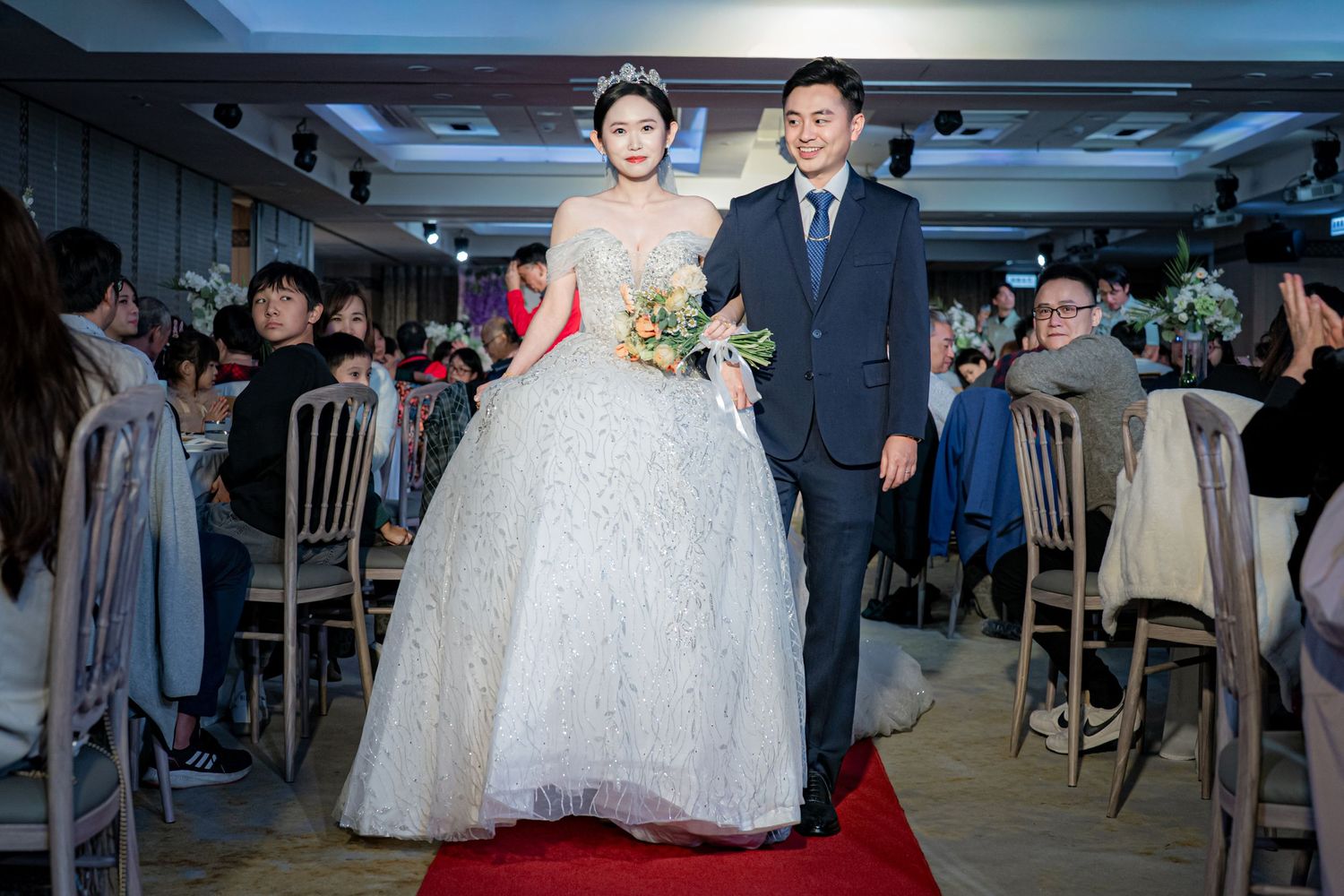 A couple walks down a red carpet aisle at a wedding reception with guests seated on both sides.