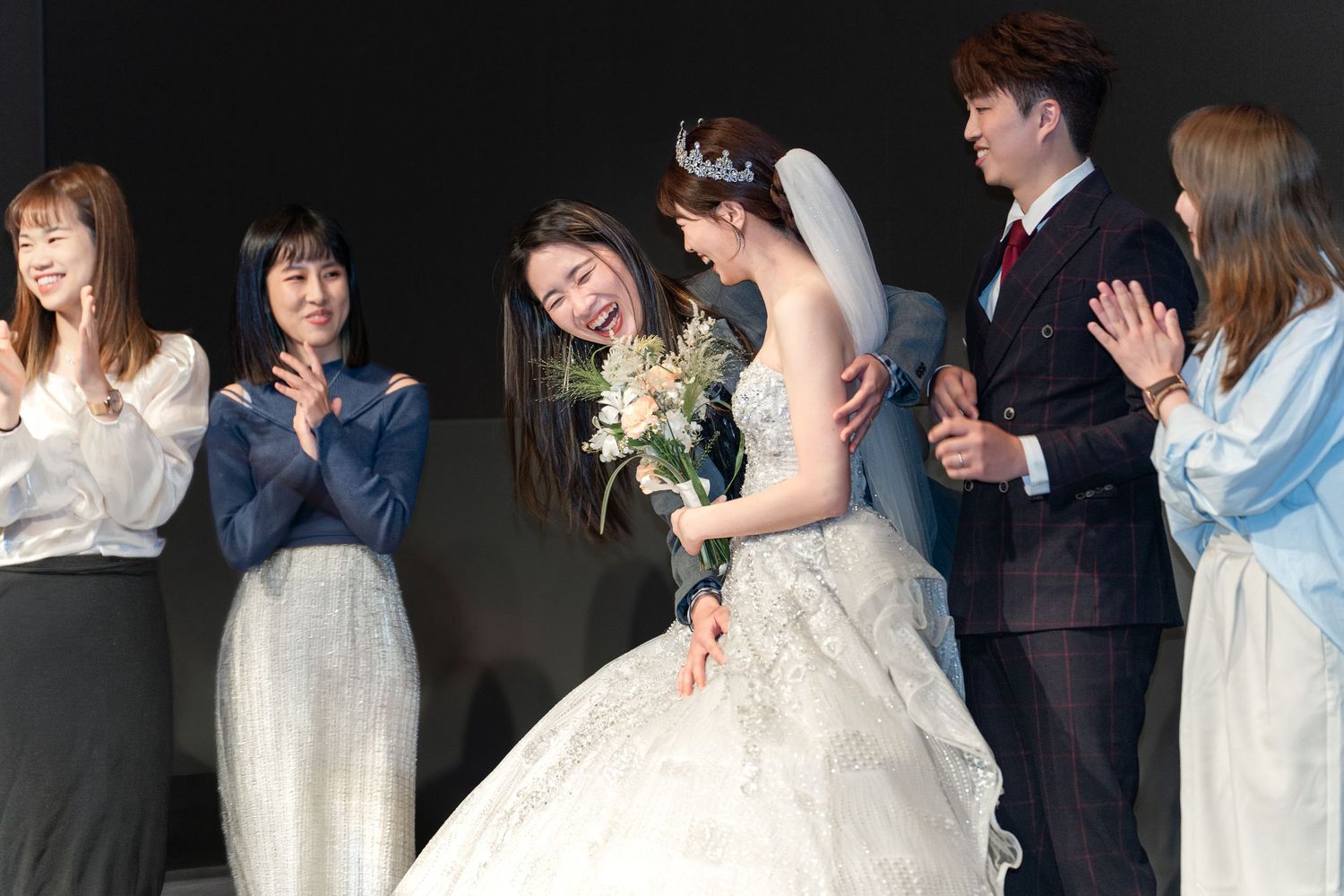 Bride in white gown with tiara holds bouquet while guests applaud at elegant wedding ceremony on stage.
